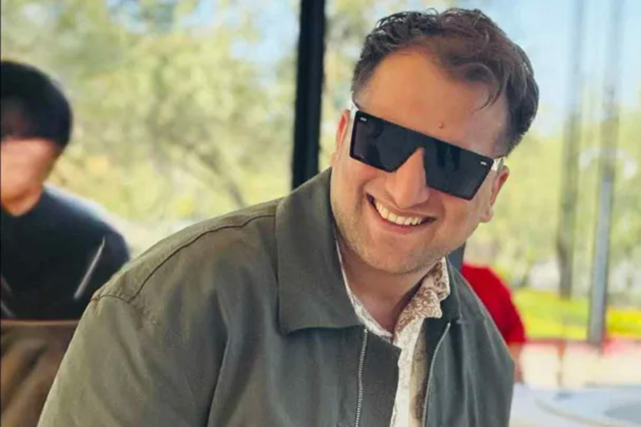 A man smiles broadly while wearing large black sunglasses and a green jacket, with a blurred outdoor background featuring trees and another person.