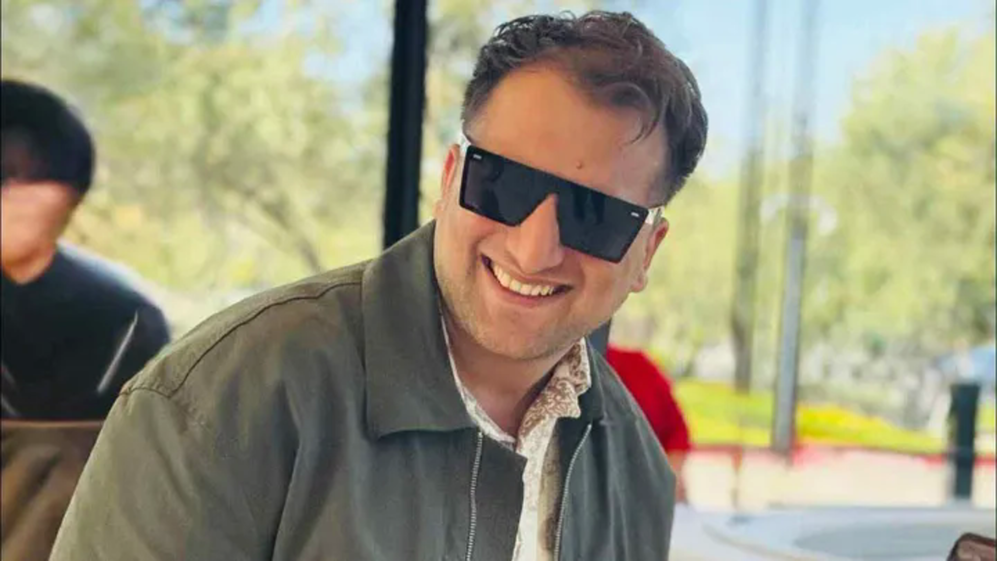A man smiles broadly while wearing large black sunglasses and a green jacket, with a blurred outdoor background featuring trees and another person.