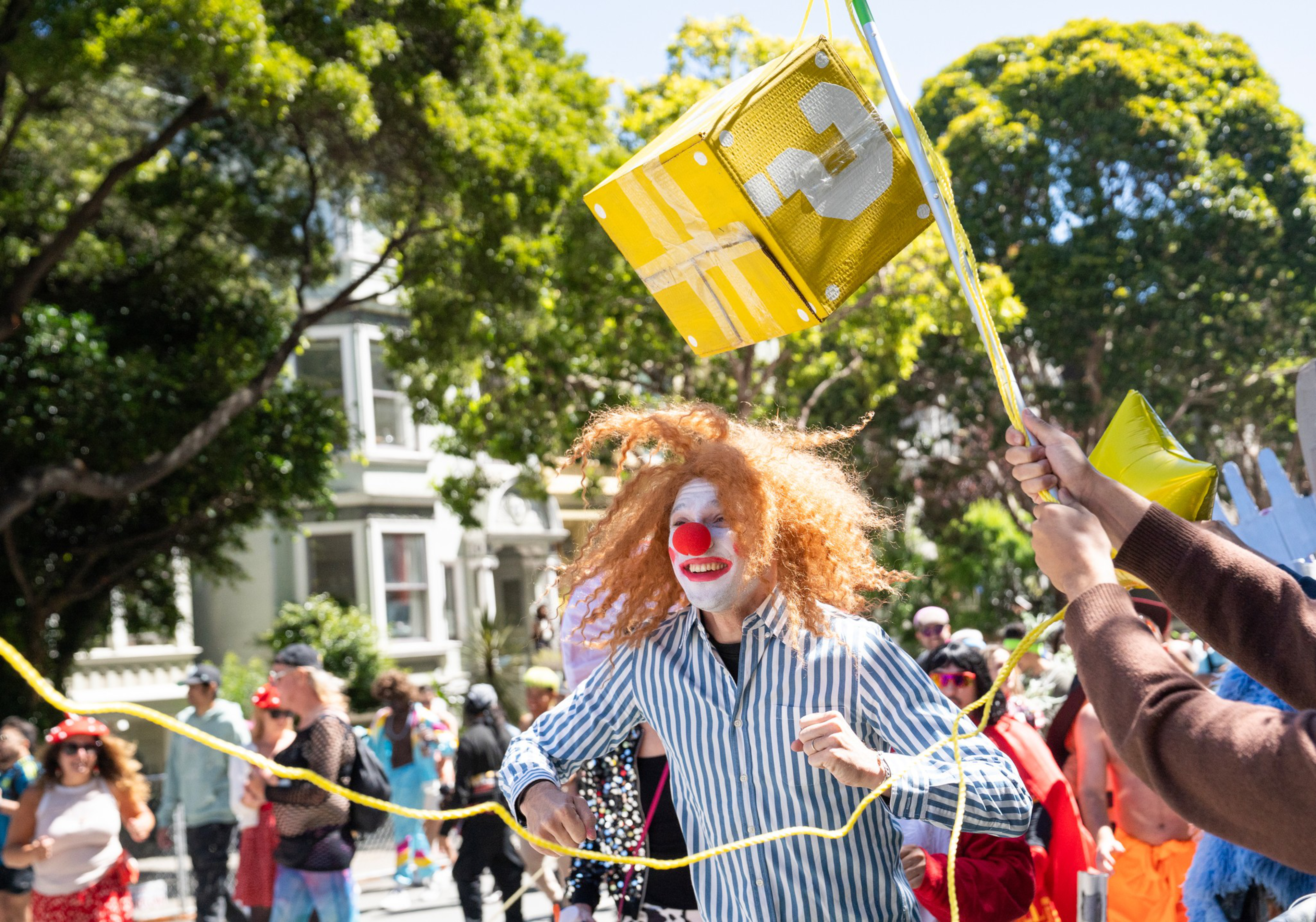 A person with clown makeup, a red nose, and curly red hair runs while holding a yellow rope, surrounded by people and trees in a sunny outdoor setting.