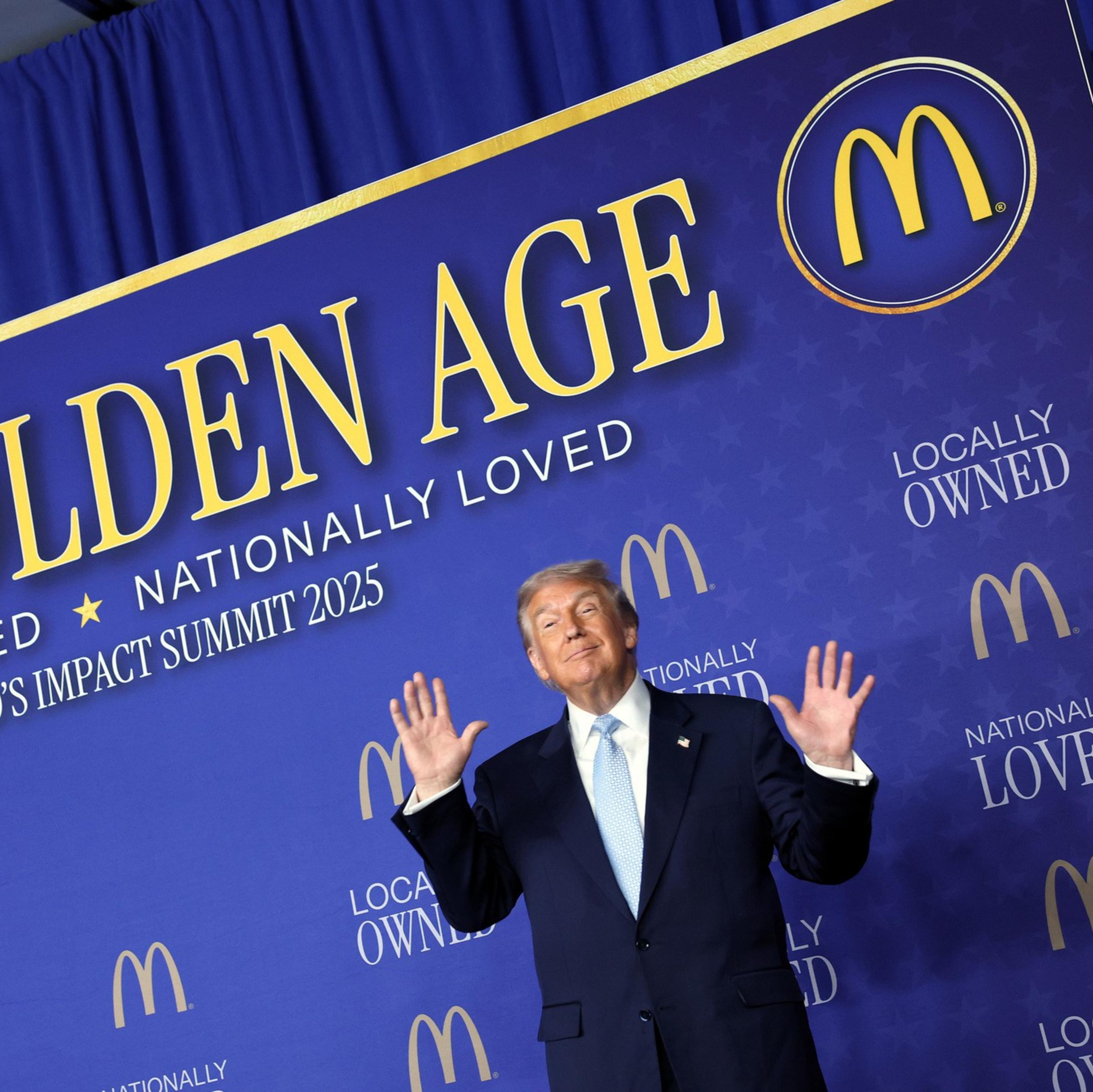 A man in a suit stands with hands raised in front of a blue backdrop that reads "THE GOLDEN AGE," "LOCALLY OWNED," and "NATIONALLY LOVED" with McDonald's logos.