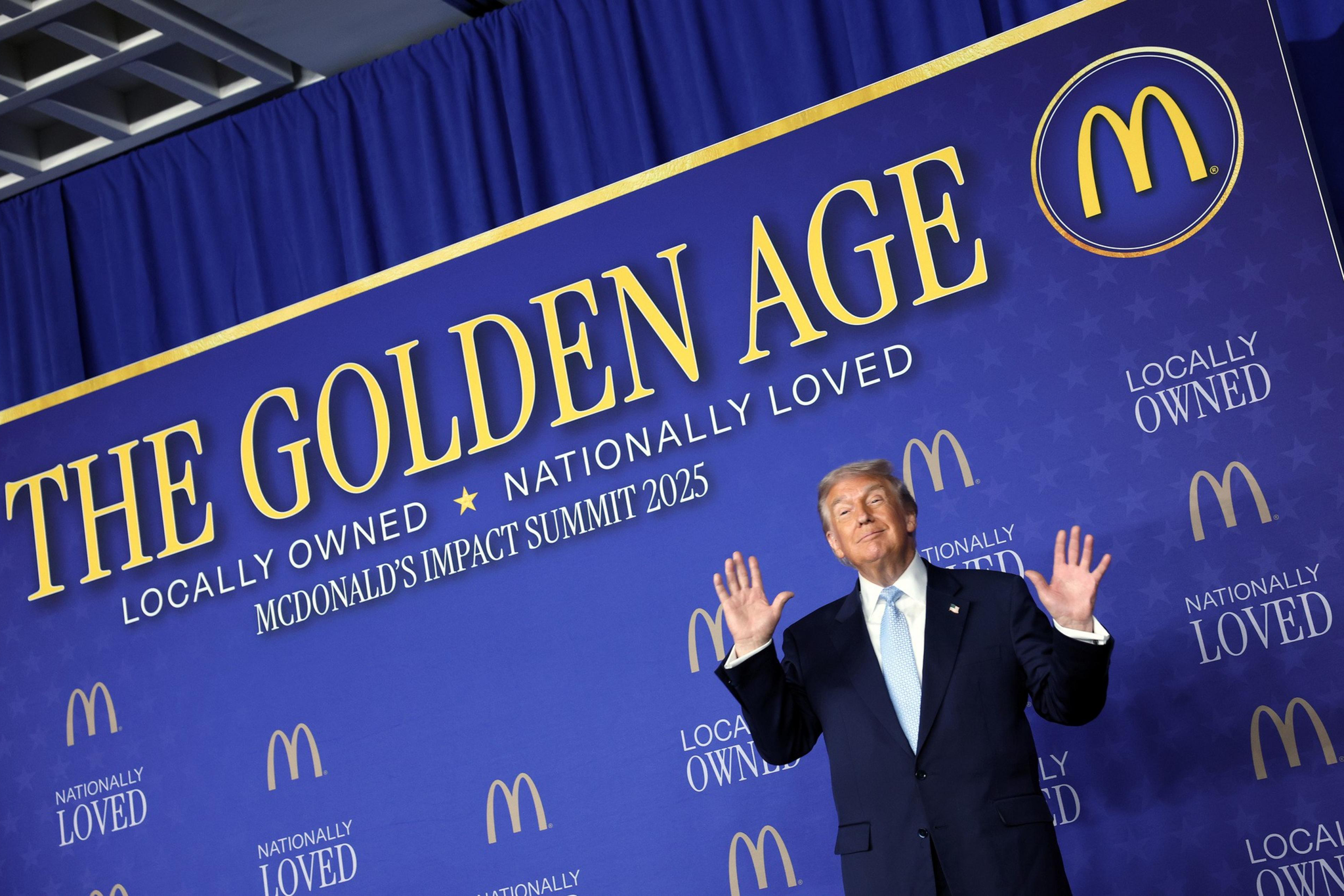 A man in a suit stands with hands raised in front of a blue backdrop that reads "THE GOLDEN AGE," "LOCALLY OWNED," and "NATIONALLY LOVED" with McDonald's logos.