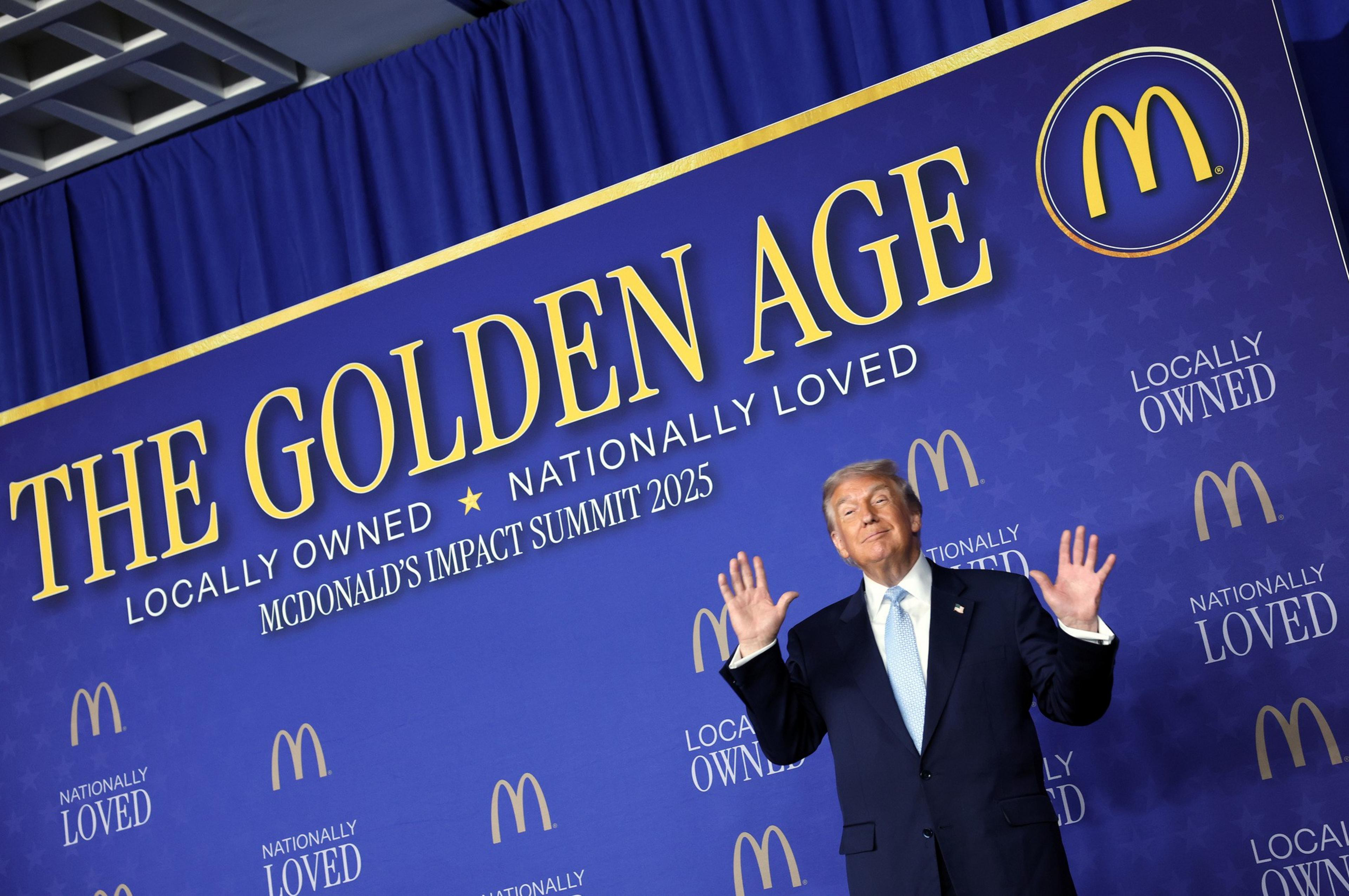 A man in a suit stands with hands raised in front of a blue backdrop that reads "THE GOLDEN AGE," "LOCALLY OWNED," and "NATIONALLY LOVED" with McDonald's logos.
