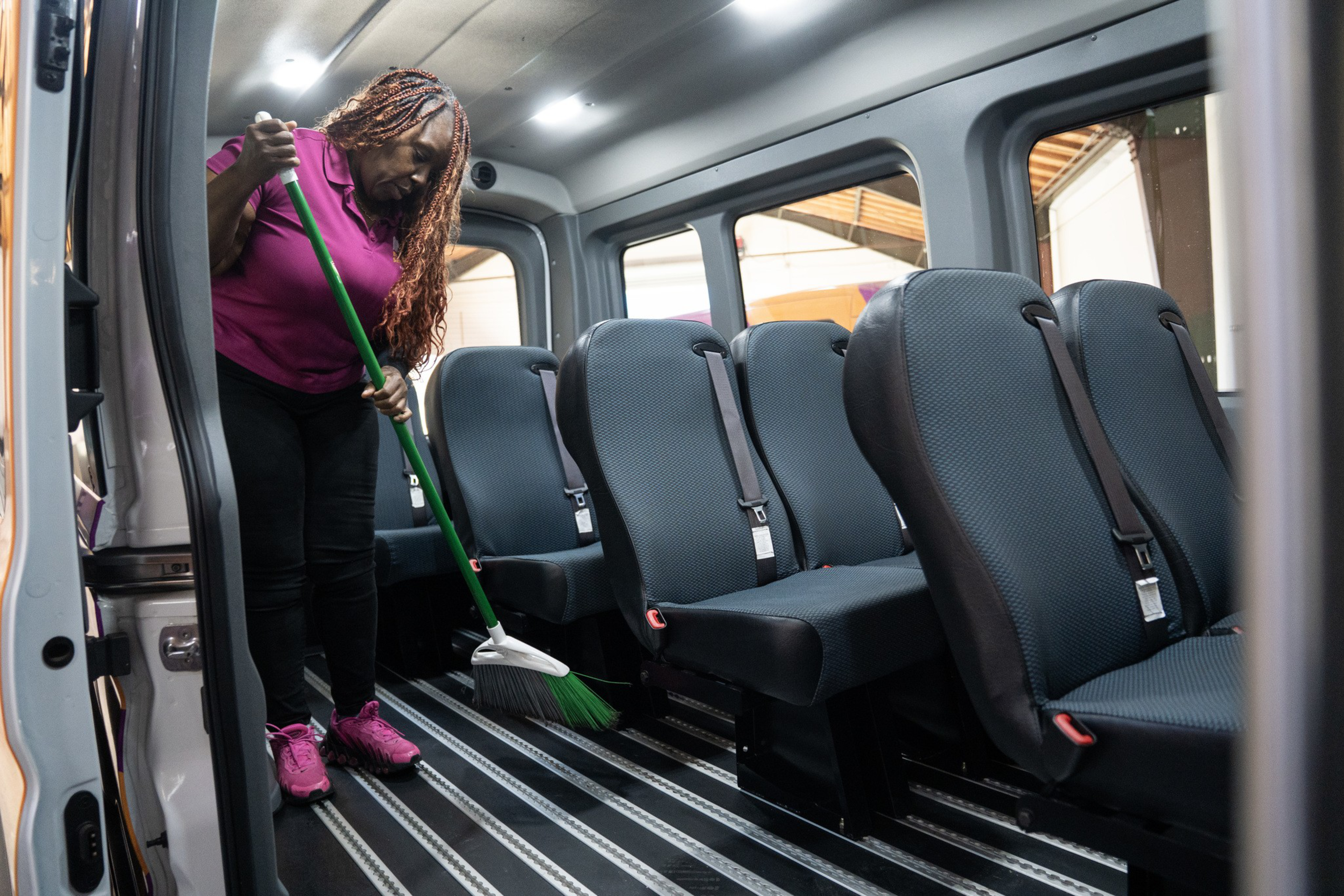 A woman with braided hair sweeps the floor inside a passenger van with black seats and seatbelts, wearing a pink shirt and matching pink shoes.