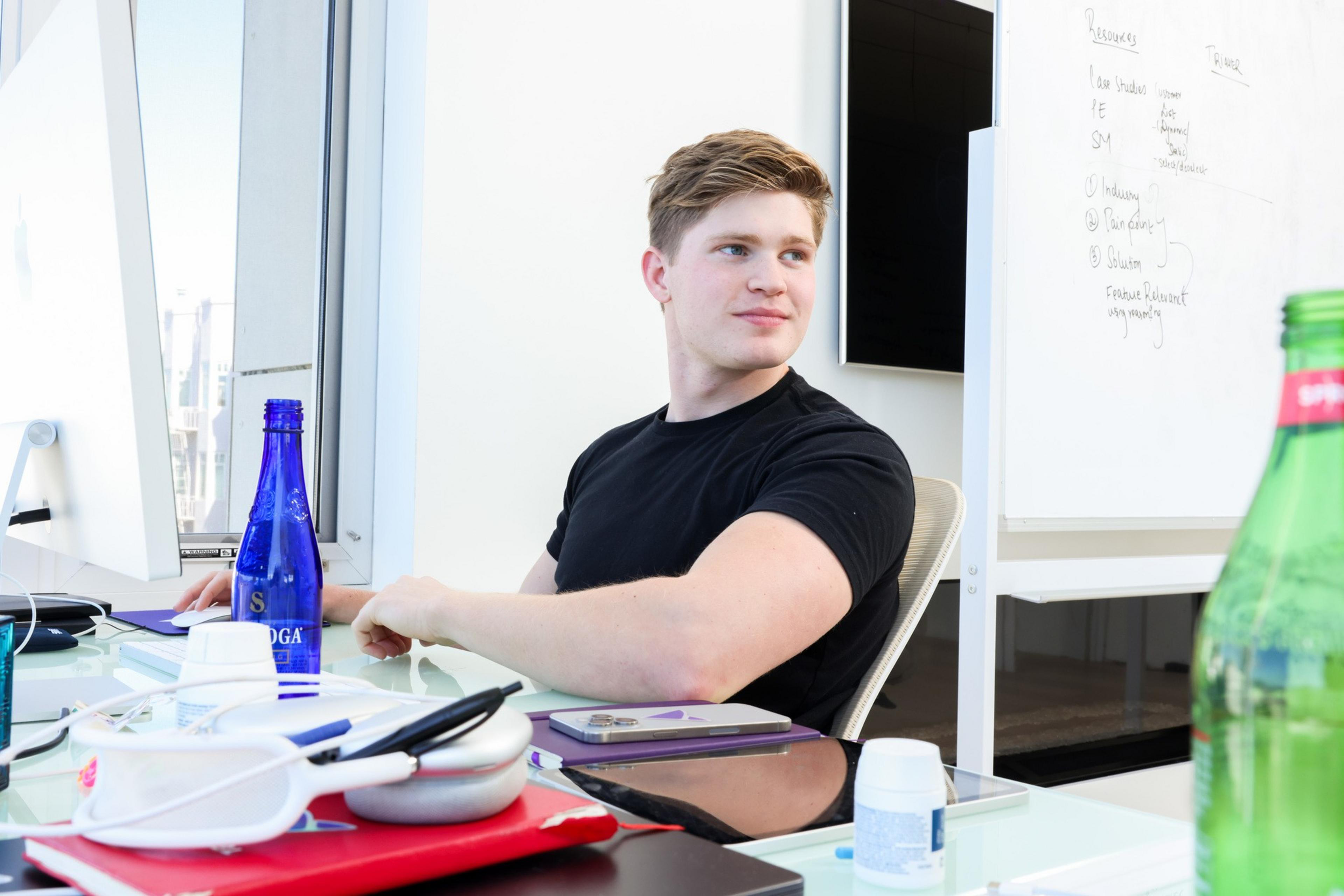 A person in a black shirt sits at a desk with a computer, looking to the side. The desk is cluttered with notebooks, bottles, pens, and a whiteboard in the background.