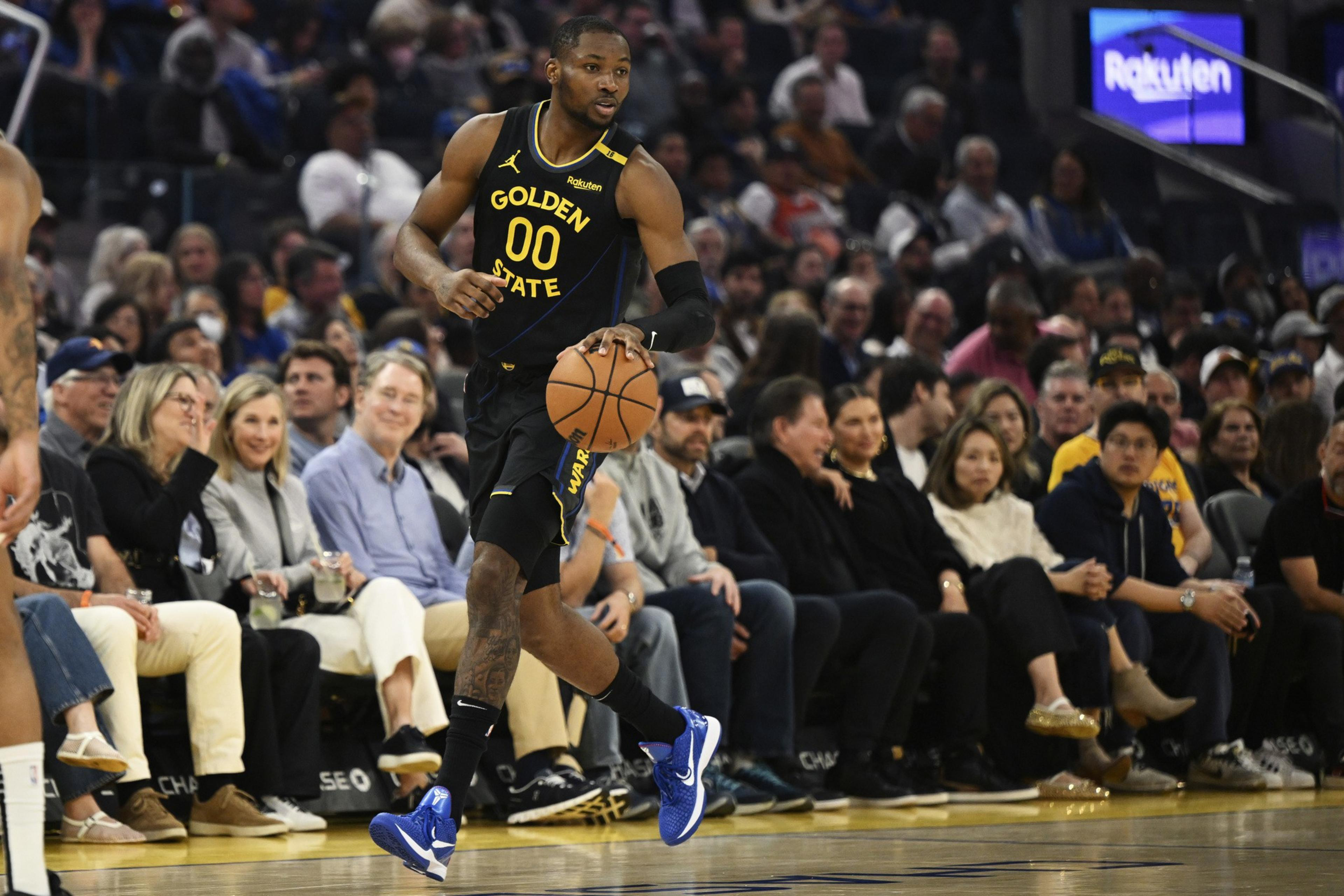 A basketball player in a Golden State uniform dribbles the ball during a game. He's wearing blue shoes and there's a crowd of spectators watching intently.