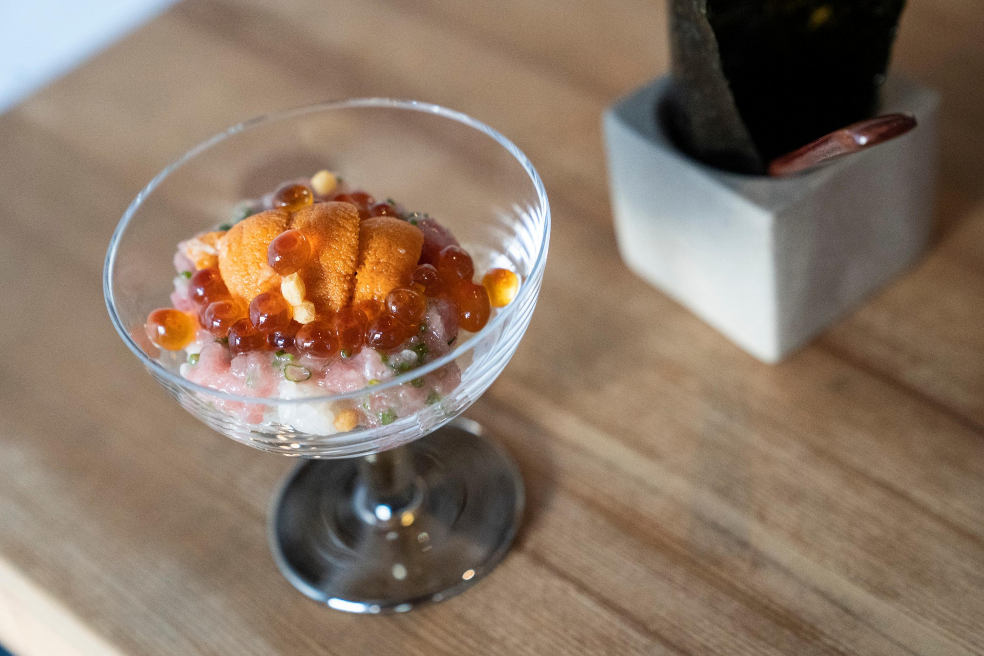 The image shows a dessert-like dish in a glass coupe, topped with orange sea urchin, red roe, and small crunchy bits, placed on a wooden surface next to a container.