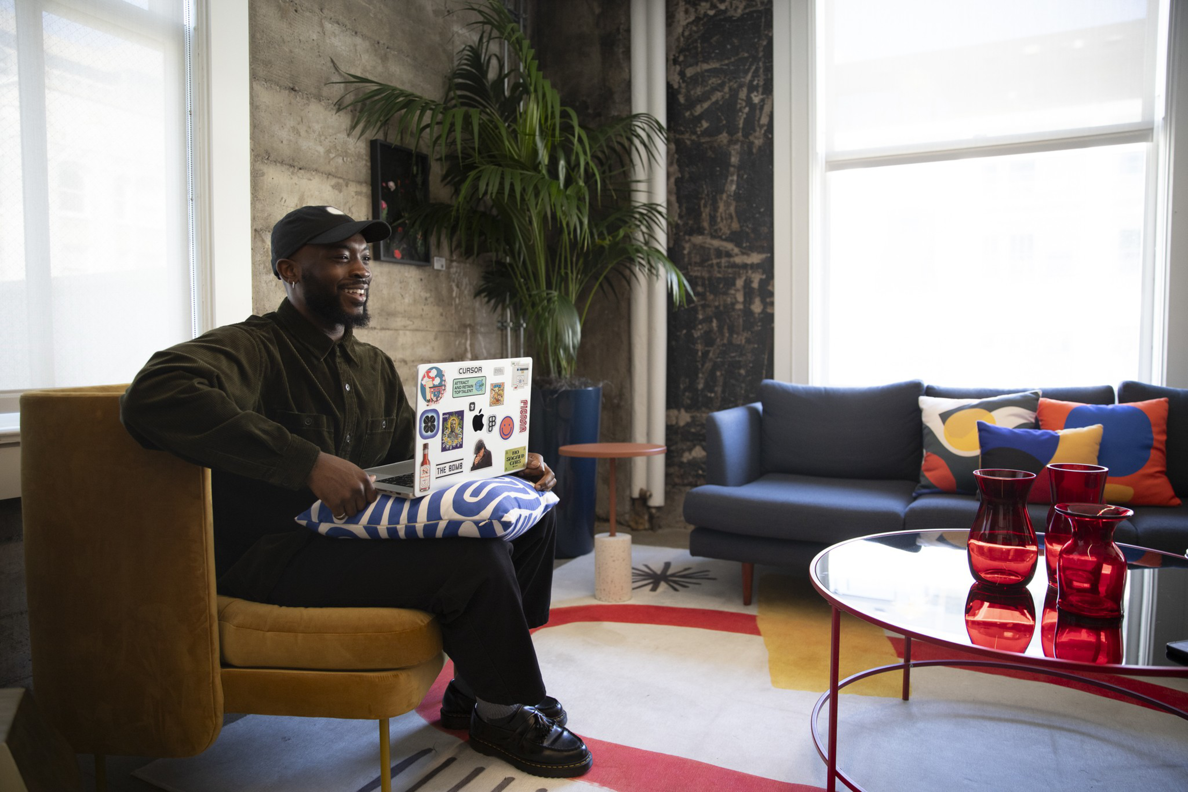 A man wearing a black cap and dark clothes sits on a mustard chair, holding a laptop on a blue-and-white pillow in a colorful, modern living room.