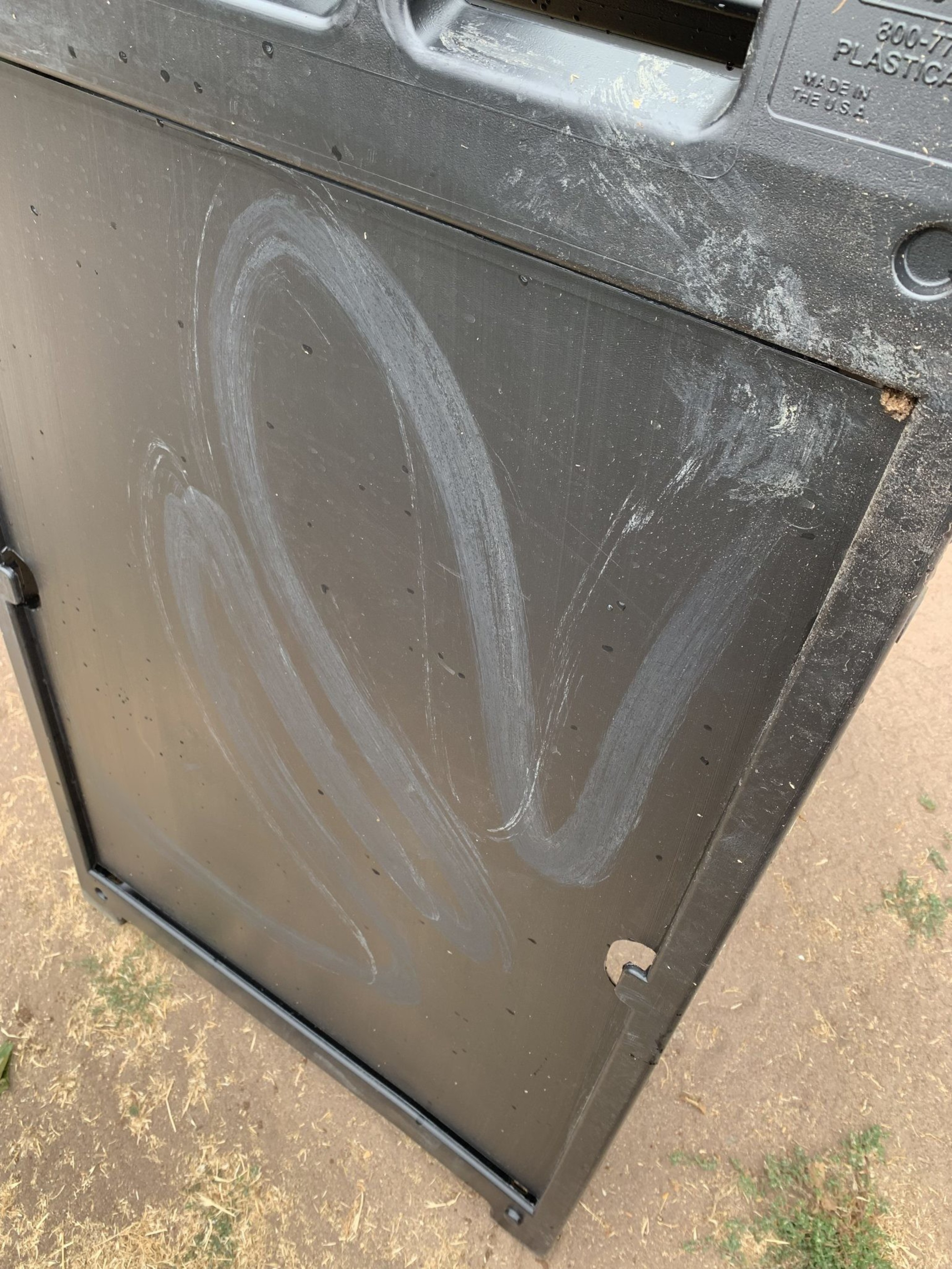 The image shows a partially opened black plastic trash can lid with dirty smudges and scratches on its surface, likely from use and exposure to the elements.