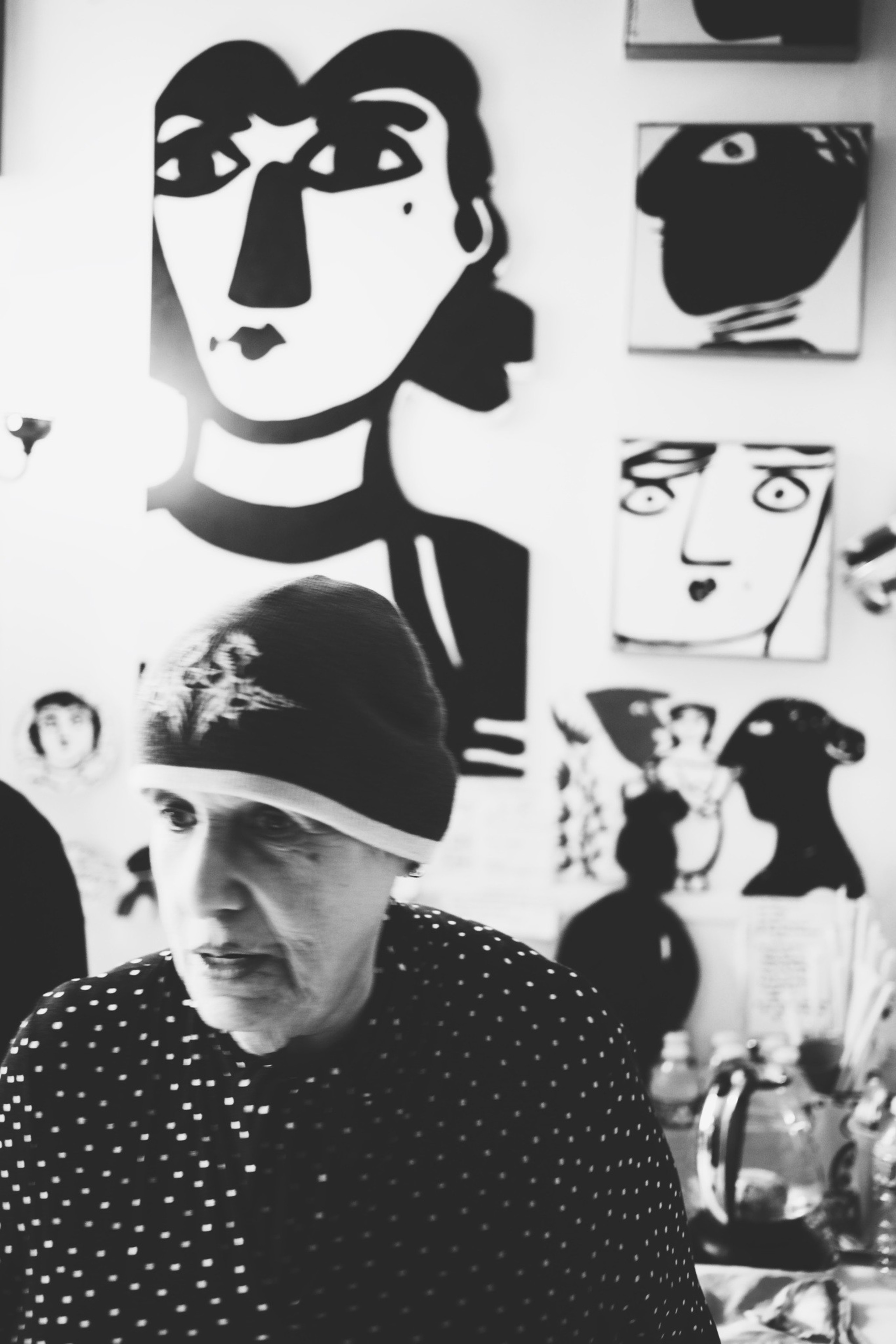 An older woman wearing a polka-dot shirt and a hat stands in front of a wall filled with bold, abstract black-and-white face artwork.