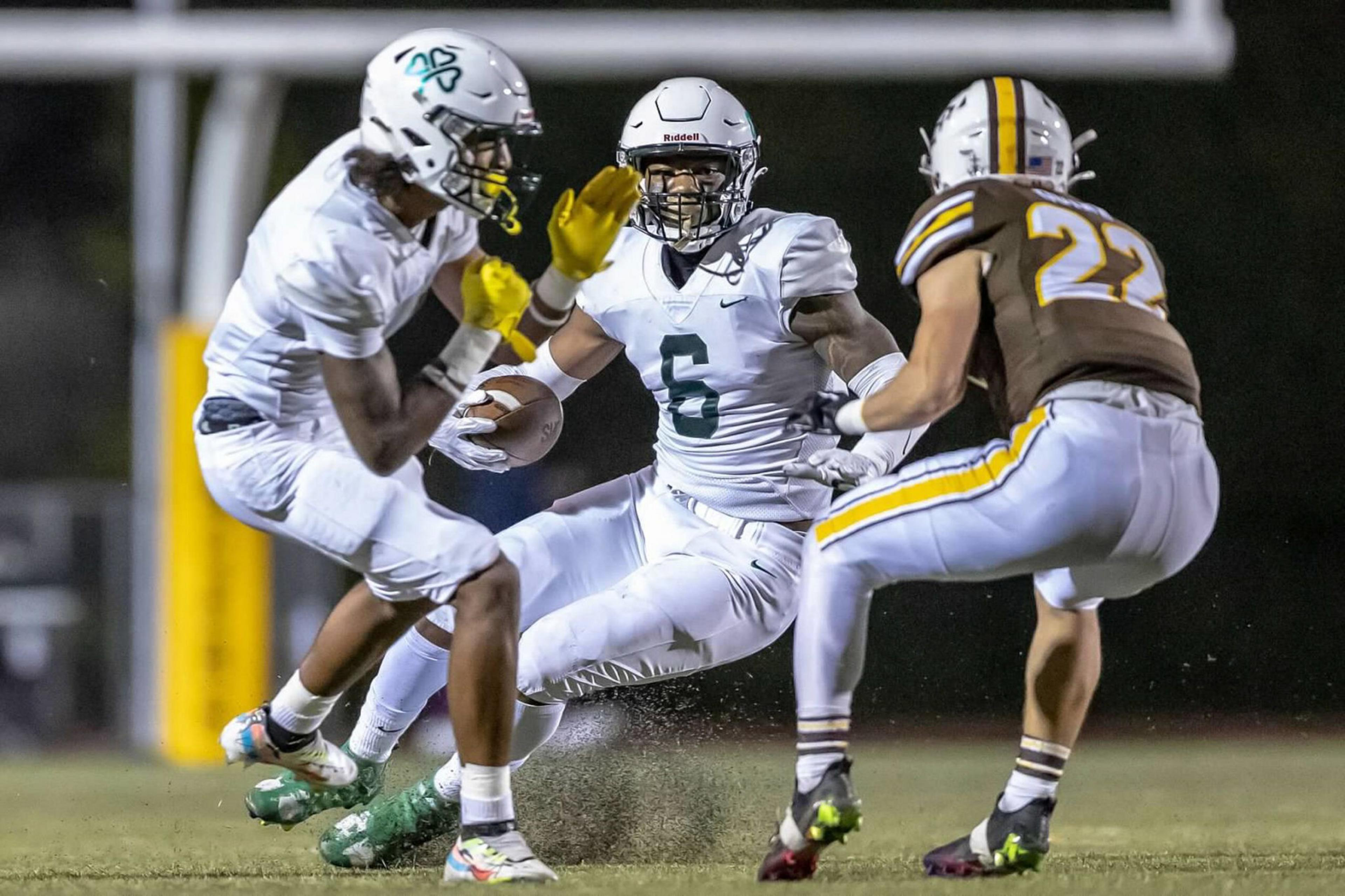 A football player in white with the ball braces as a teammate blocks while a defender in brown and yellow approaches during a night game.