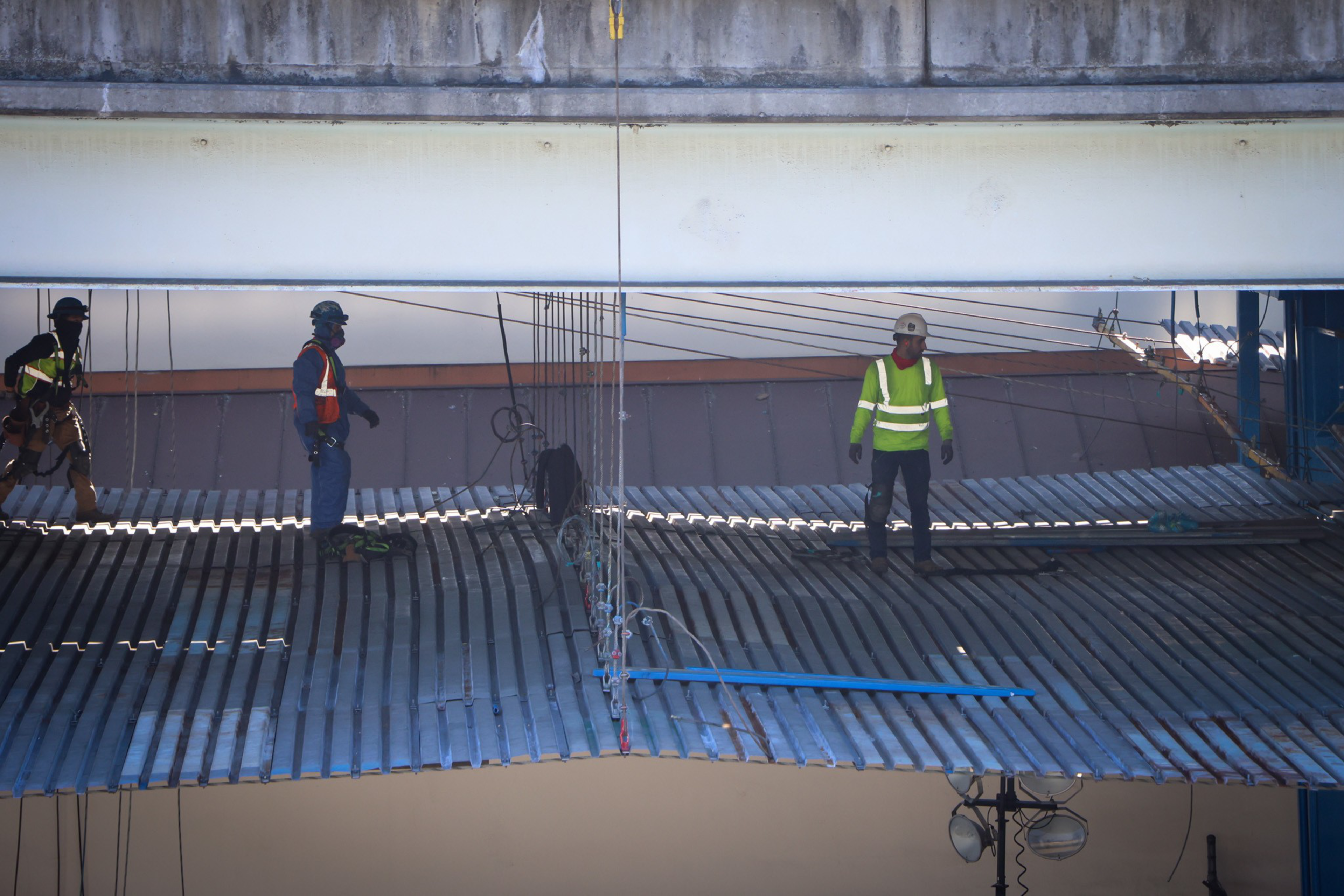 Three construction workers wearing safety gear stand on metal beams under a bridge, engaged in ongoing structural work.