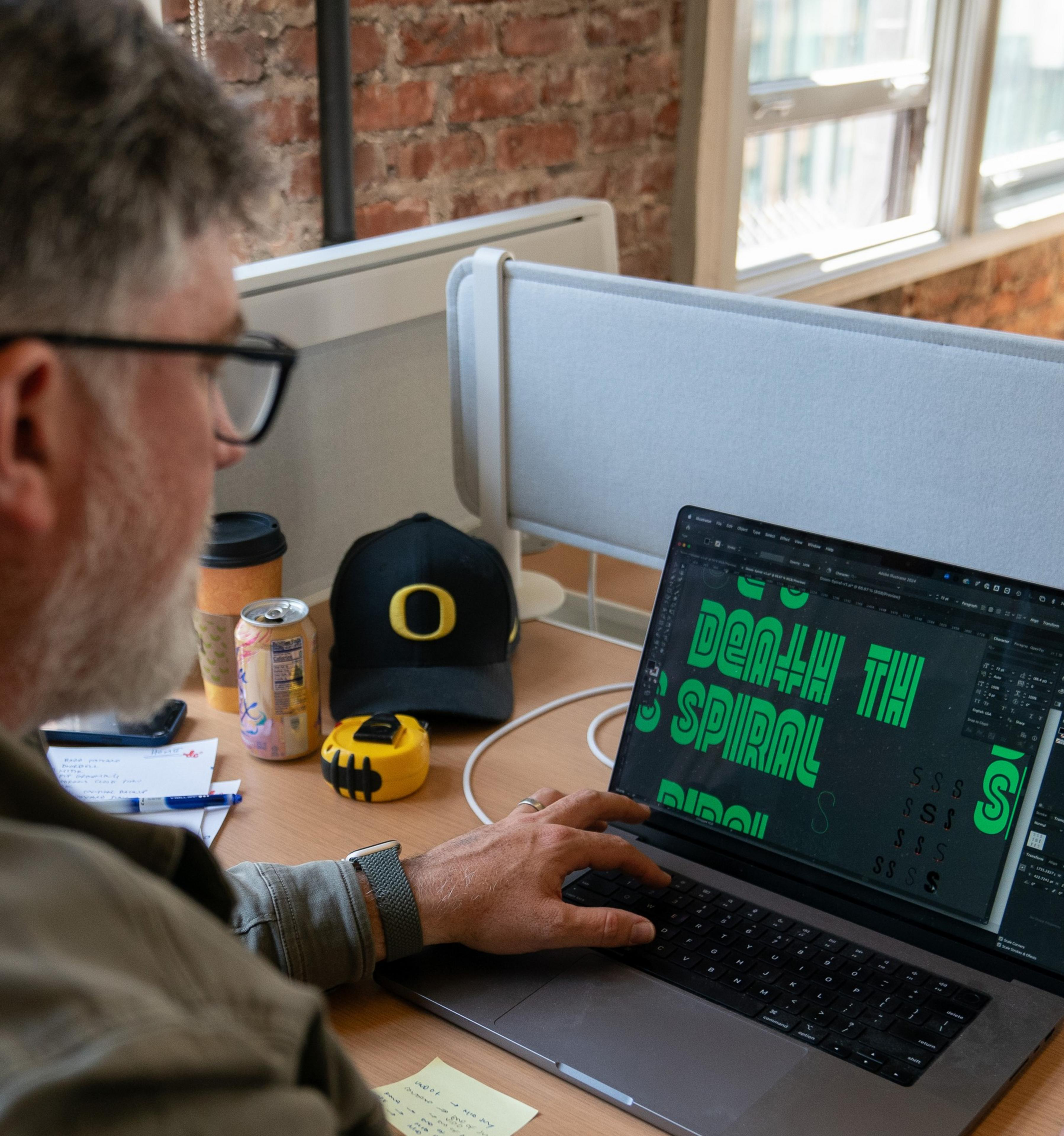 A person with glasses is working on a laptop displaying graphic design. A coffee cup, cap, soda can, and tape measure are on the desk near a window.