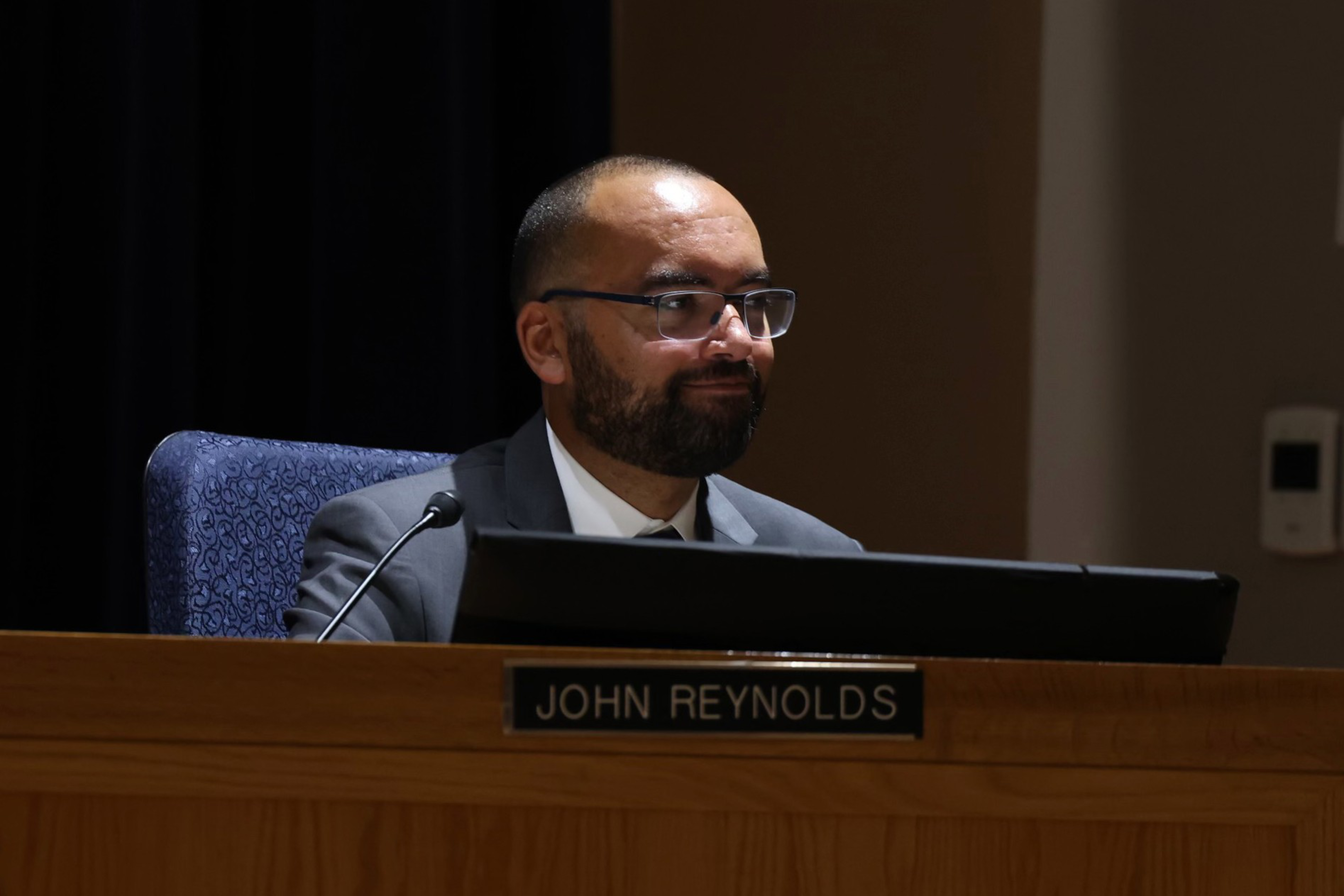A man with glasses, a beard, and a suit sits behind a wooden desk with a nameplate reading "John Reynolds."