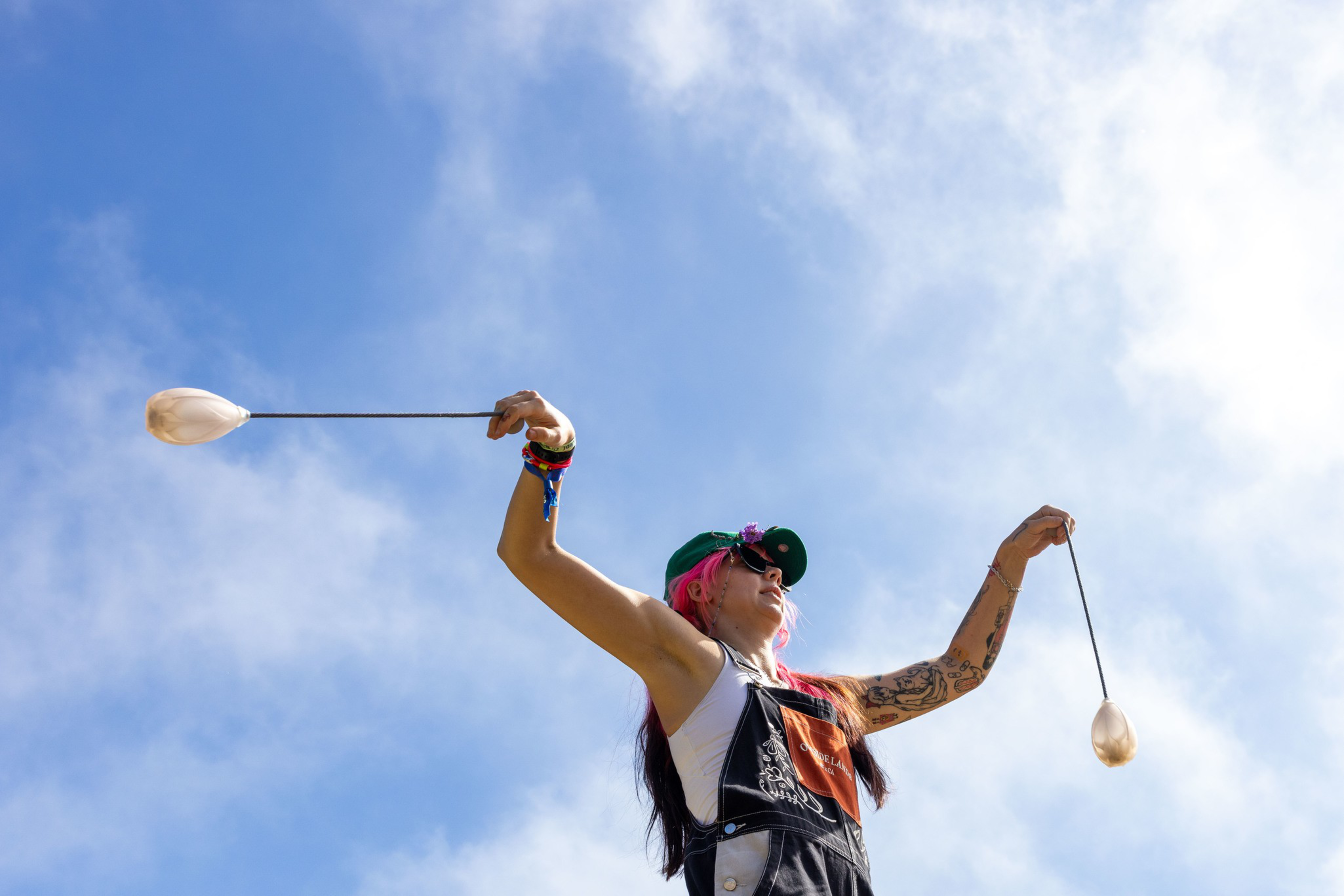 A person with pink hair and tattoos spins two poi against a blue sky, wearing sunglasses, a green cap, and a black apron over a white sleeveless top.