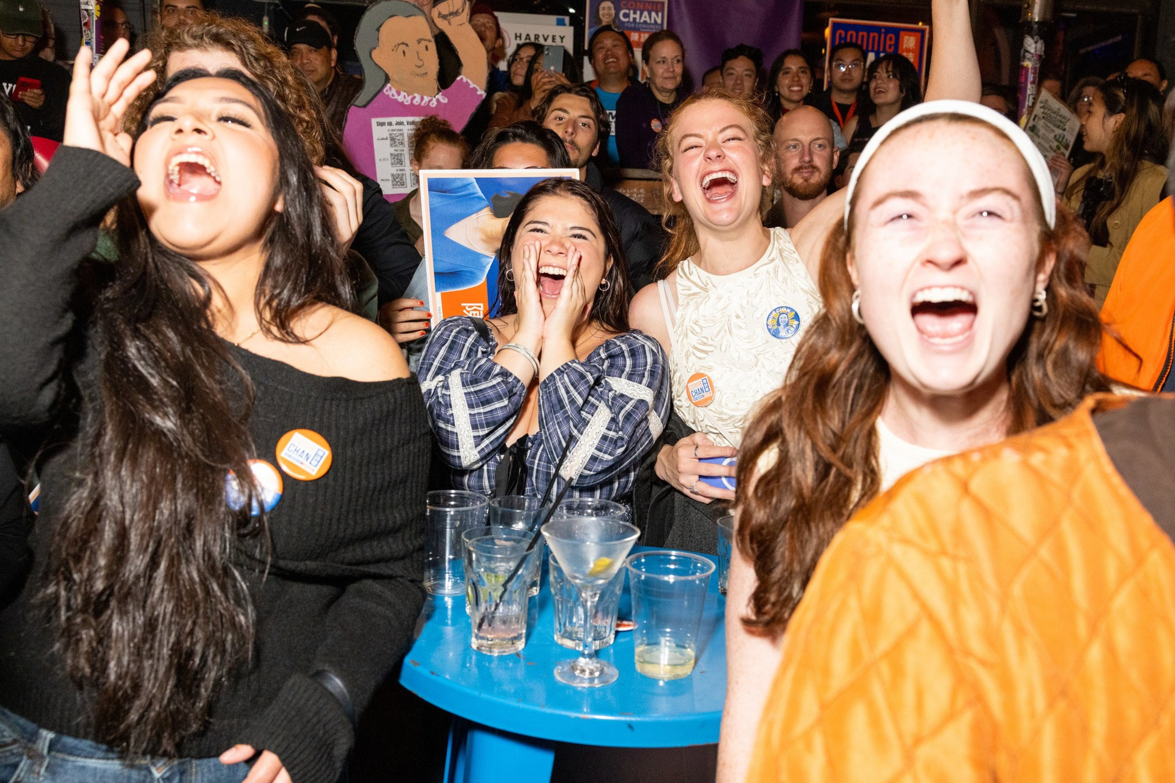 A diverse group of people cheer loudly, smiling and laughing, around a blue table with drinks at a crowded, lively event.