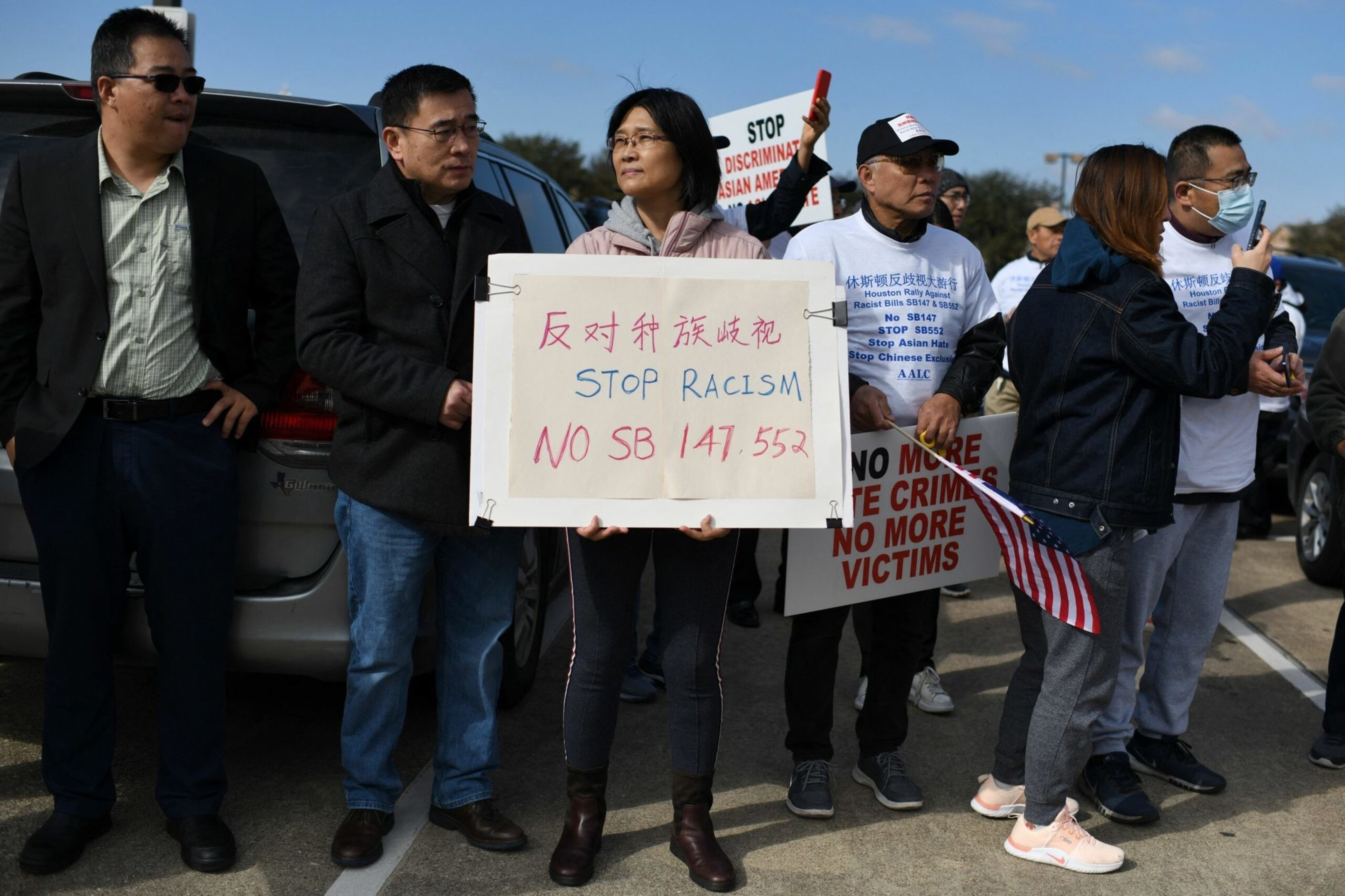 A group of people holding signs protesting racism, with messages like "STOP RACISM" and "NO MORE HATE CRIMES NO MORE VICTIMS" in an outdoor setting.