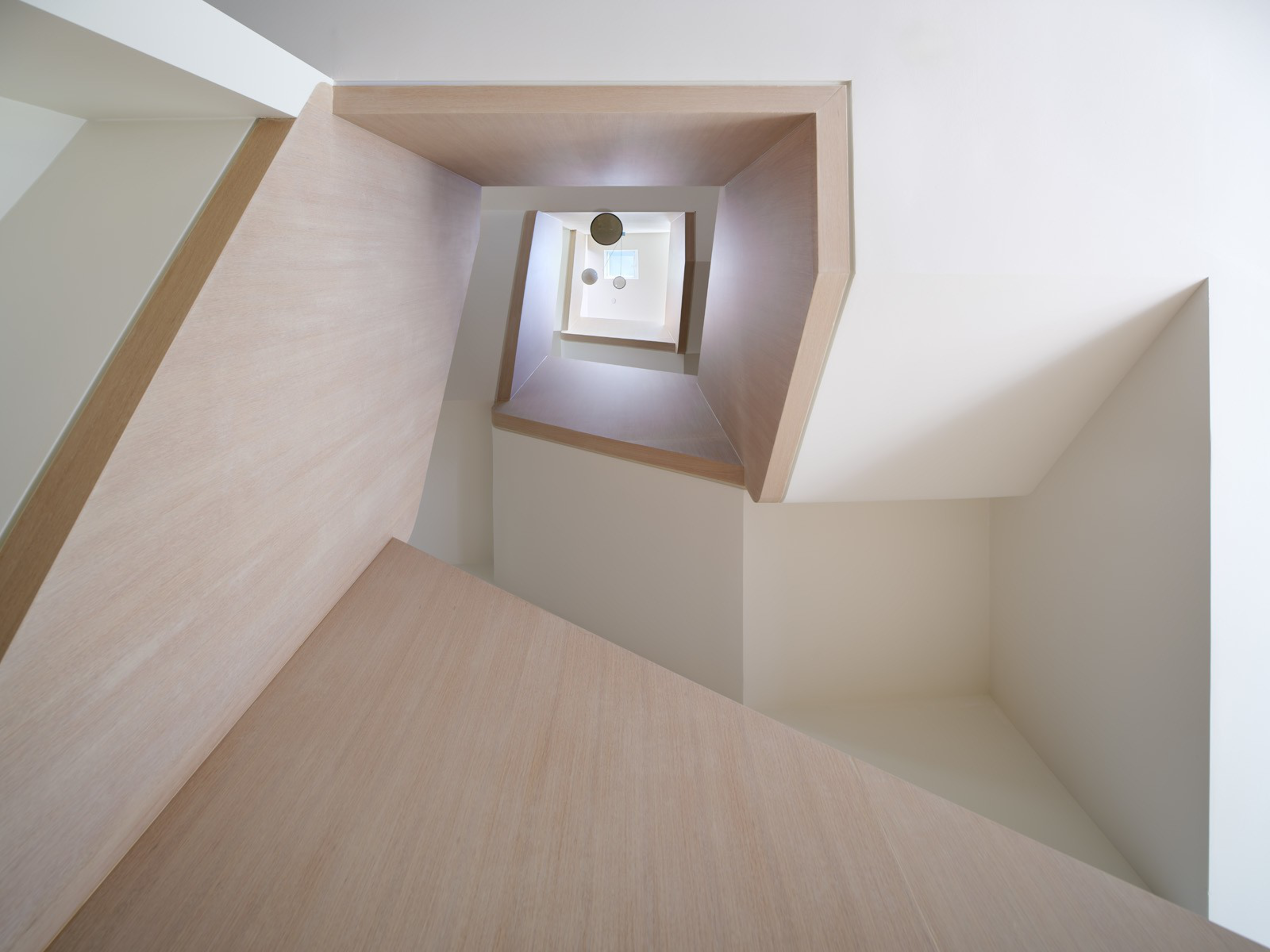 A minimalist, modern spiral staircase with beige wooden railings and white walls spirals upward, ending in a skylight with hanging spherical lights.