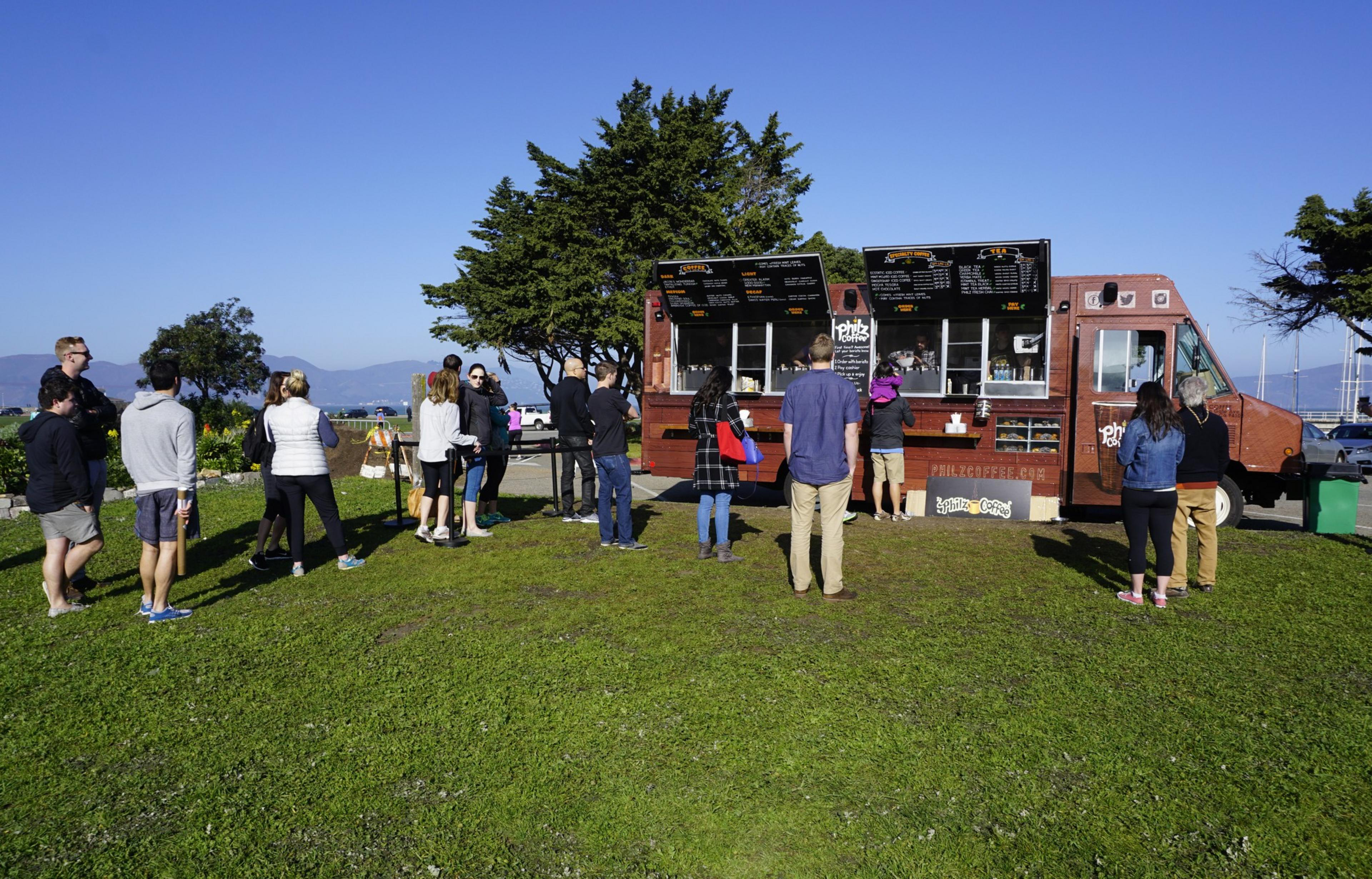 People stand in a spaced line on green grass, waiting to order at a brown food truck under clear blue skies with trees and mountains in the background.