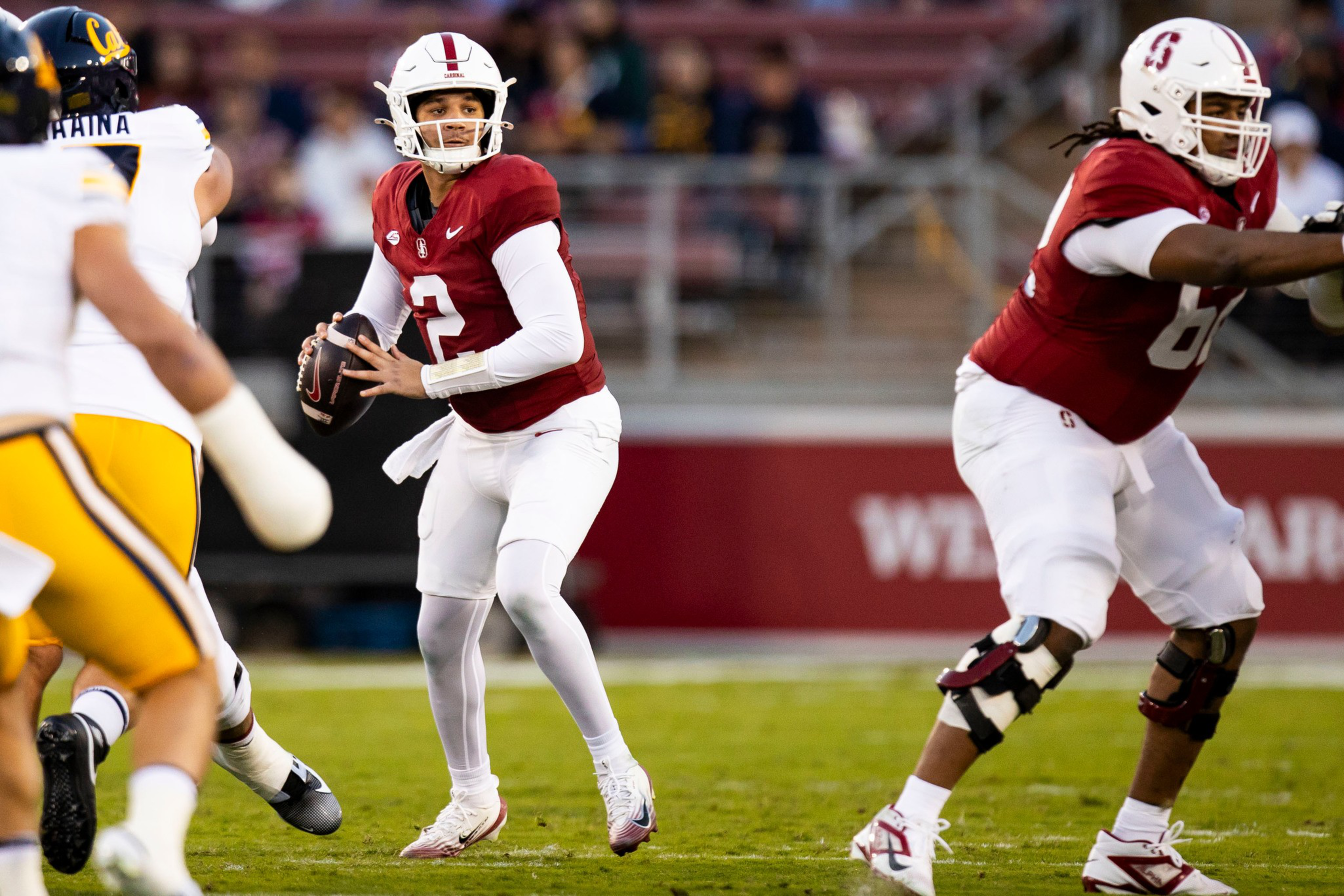 A football quarterback in a red jersey prepares to throw while a teammate blocks players in white and yellow uniforms on the field.