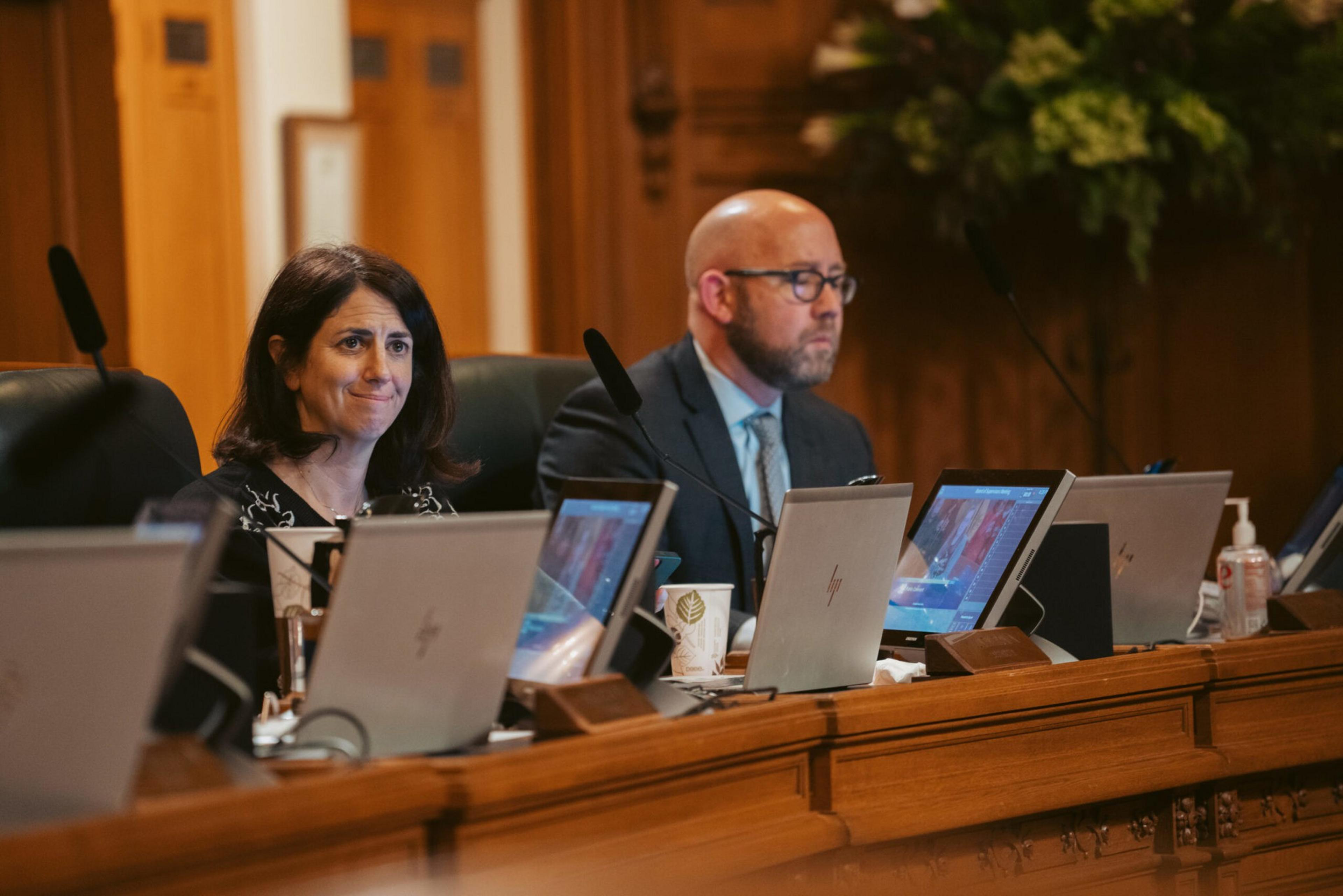 A woman and a man sit at a wood-paneled conference table with laptops, microphones, and papers in front of them during a meeting.