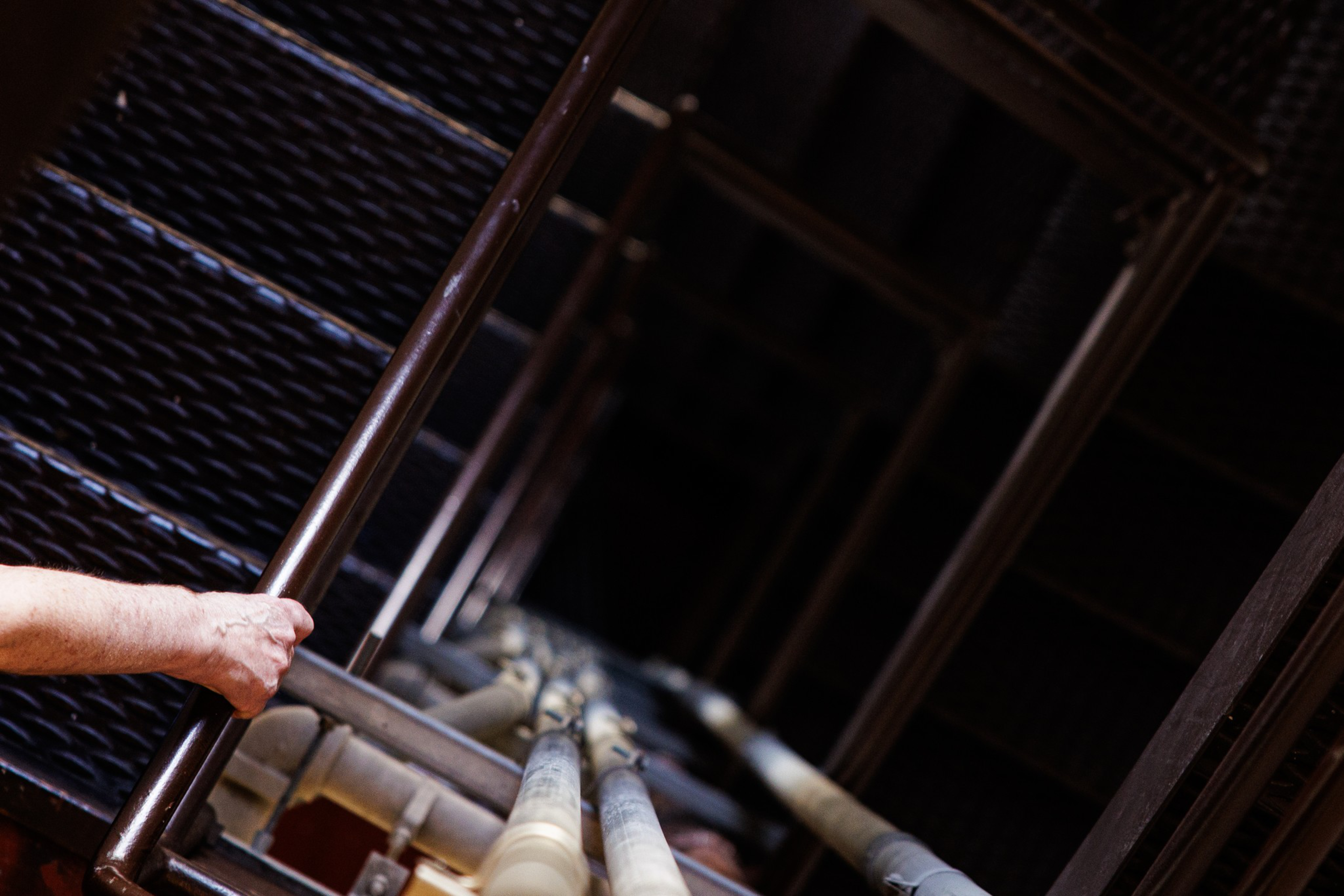 A person’s hand grasps a metal railing while descending a steep, dark industrial stairwell with exposed pipes running alongside.