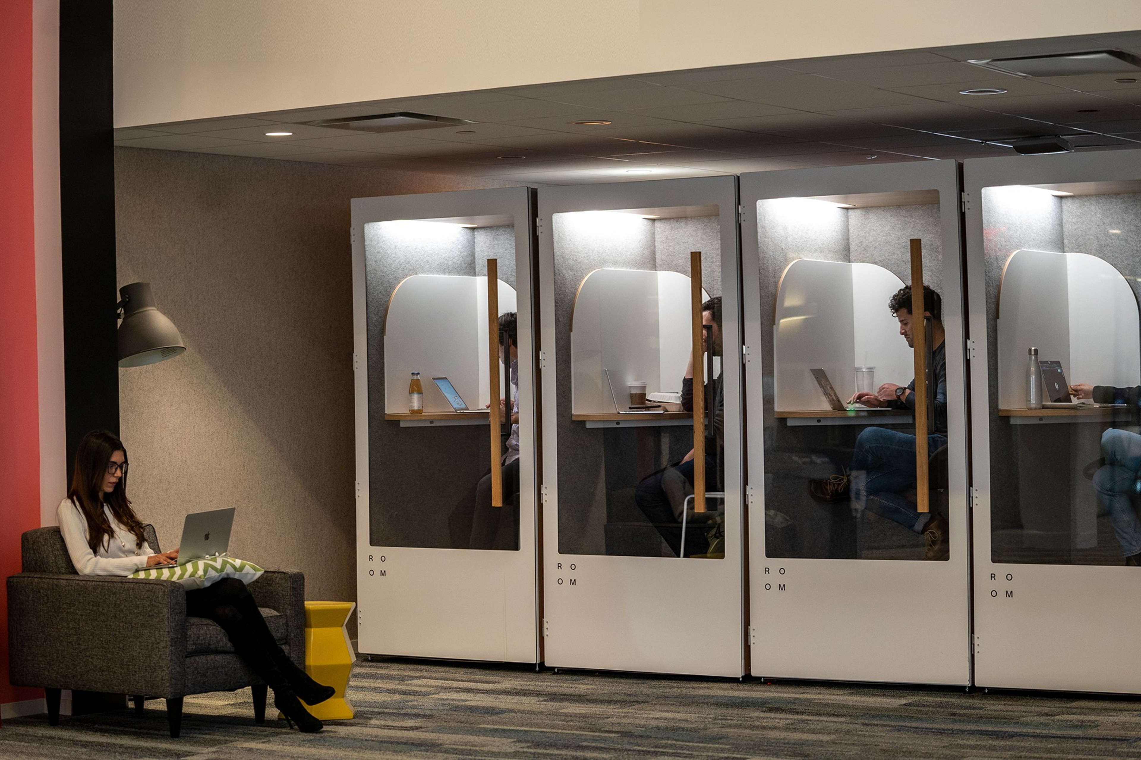 A person sits on a gray armchair with a laptop, while four individuals work in glass cubicles with laptops, each compartment well-lit and containing a small desk.