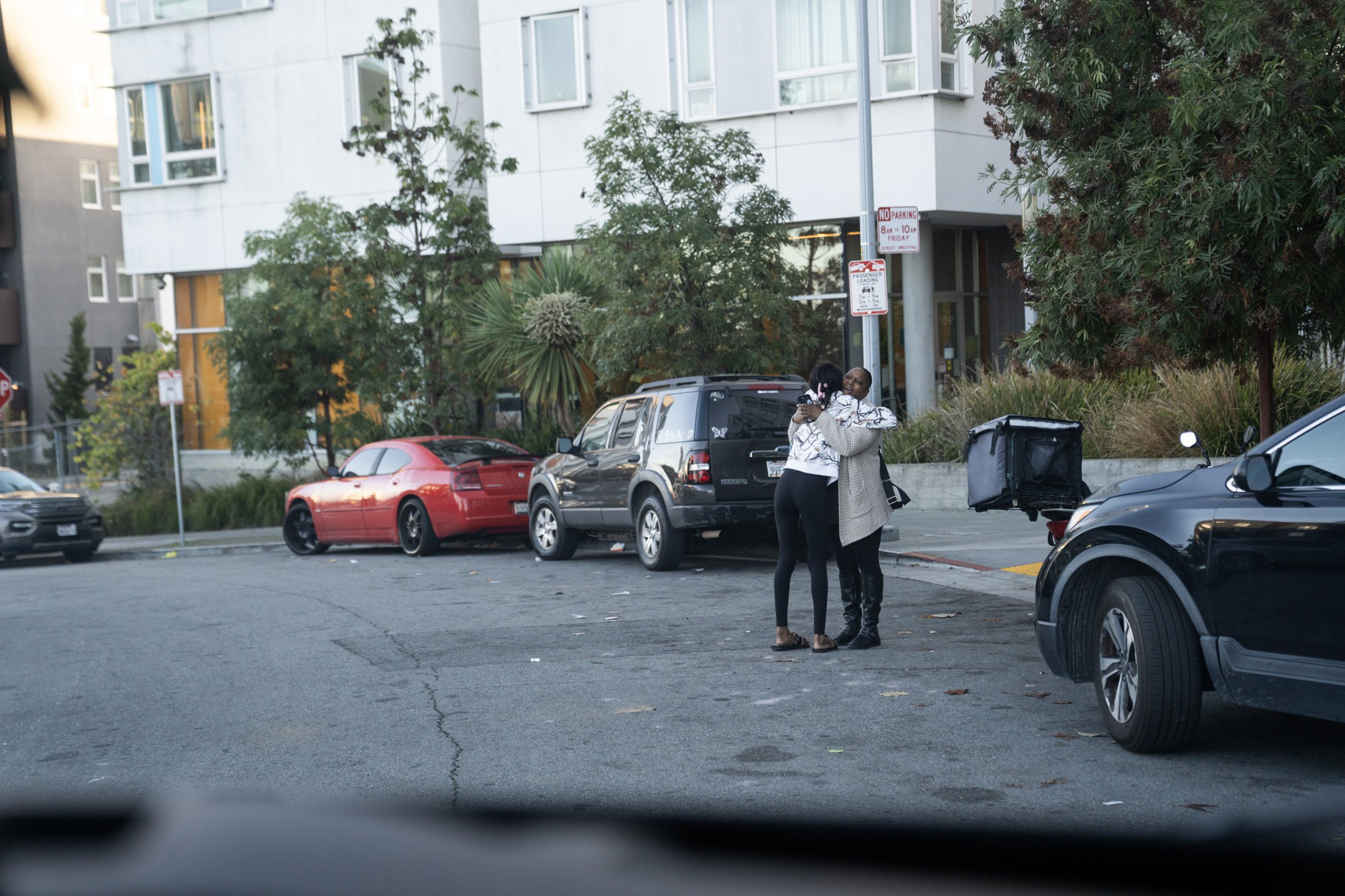 Two people stand close together, hugging on a quiet street corner with parked cars and apartment buildings in the background.