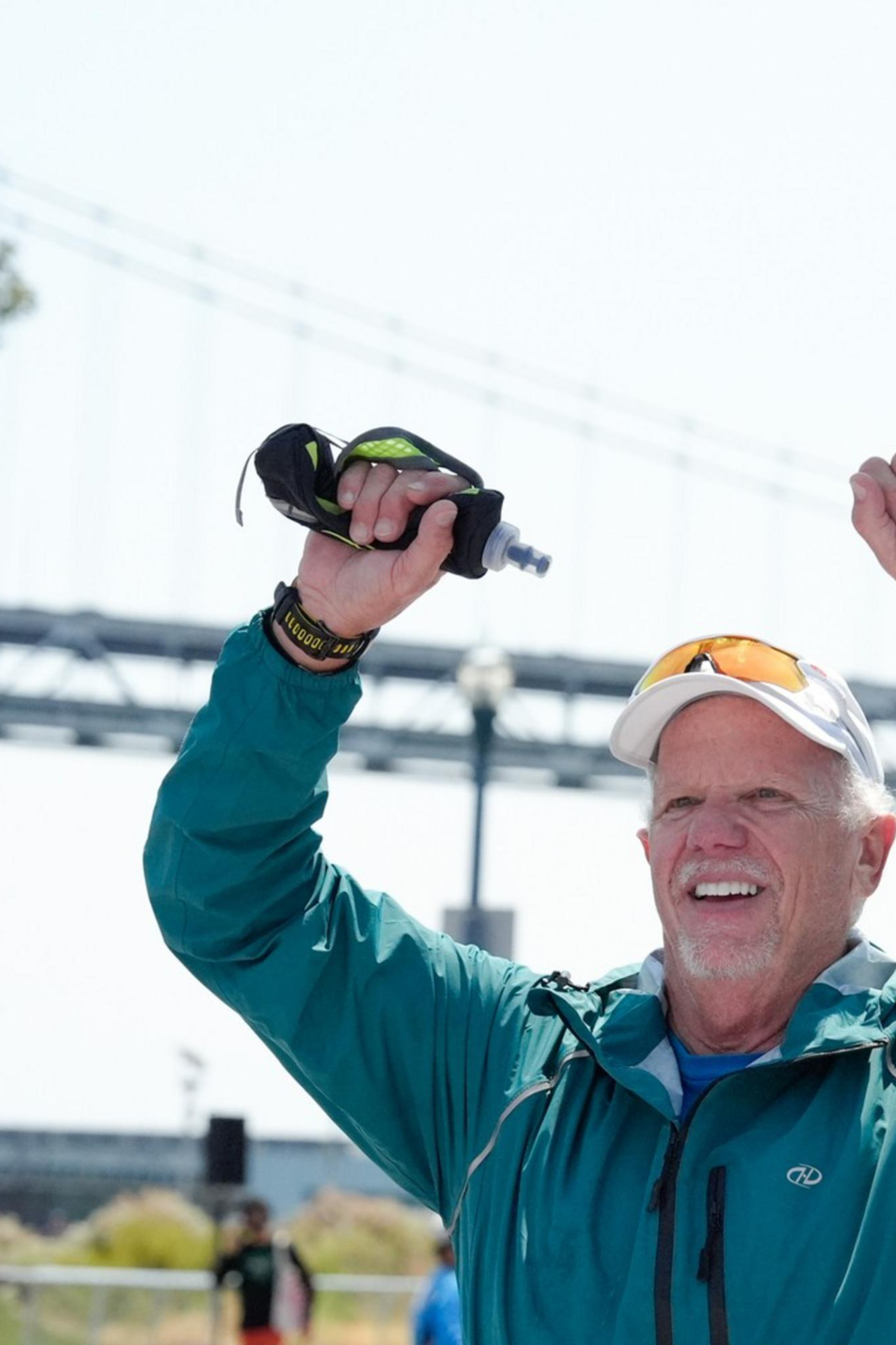 A man in a teal jacket and white cap raises his arms in victory at a marathon finish line, with a bridge and arrow sculpture in the background.