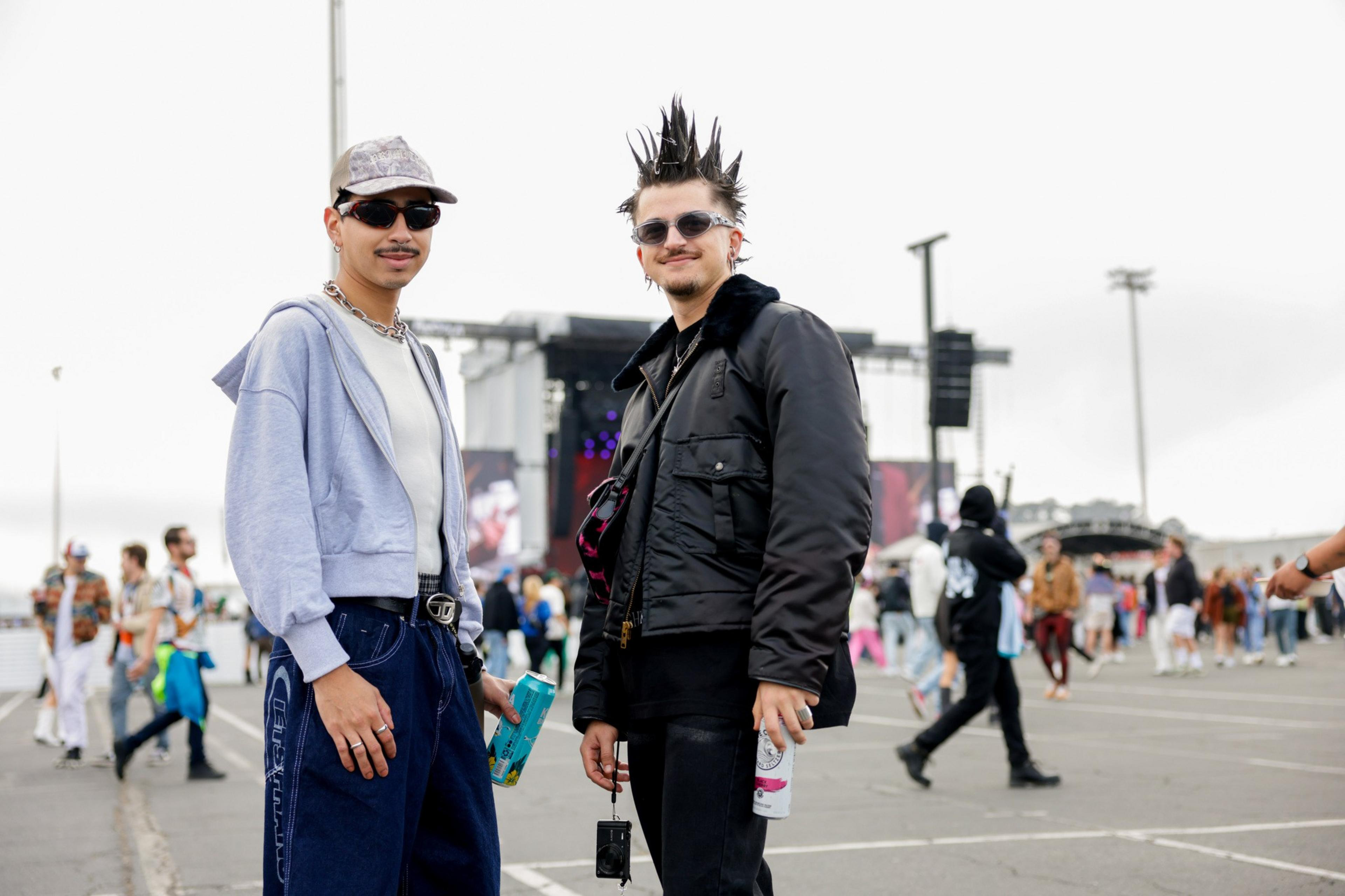 Two young men stand outside at a crowded event; one wears a light hoodie and cap, the other sports spiked hair and a black jacket, both wearing sunglasses.