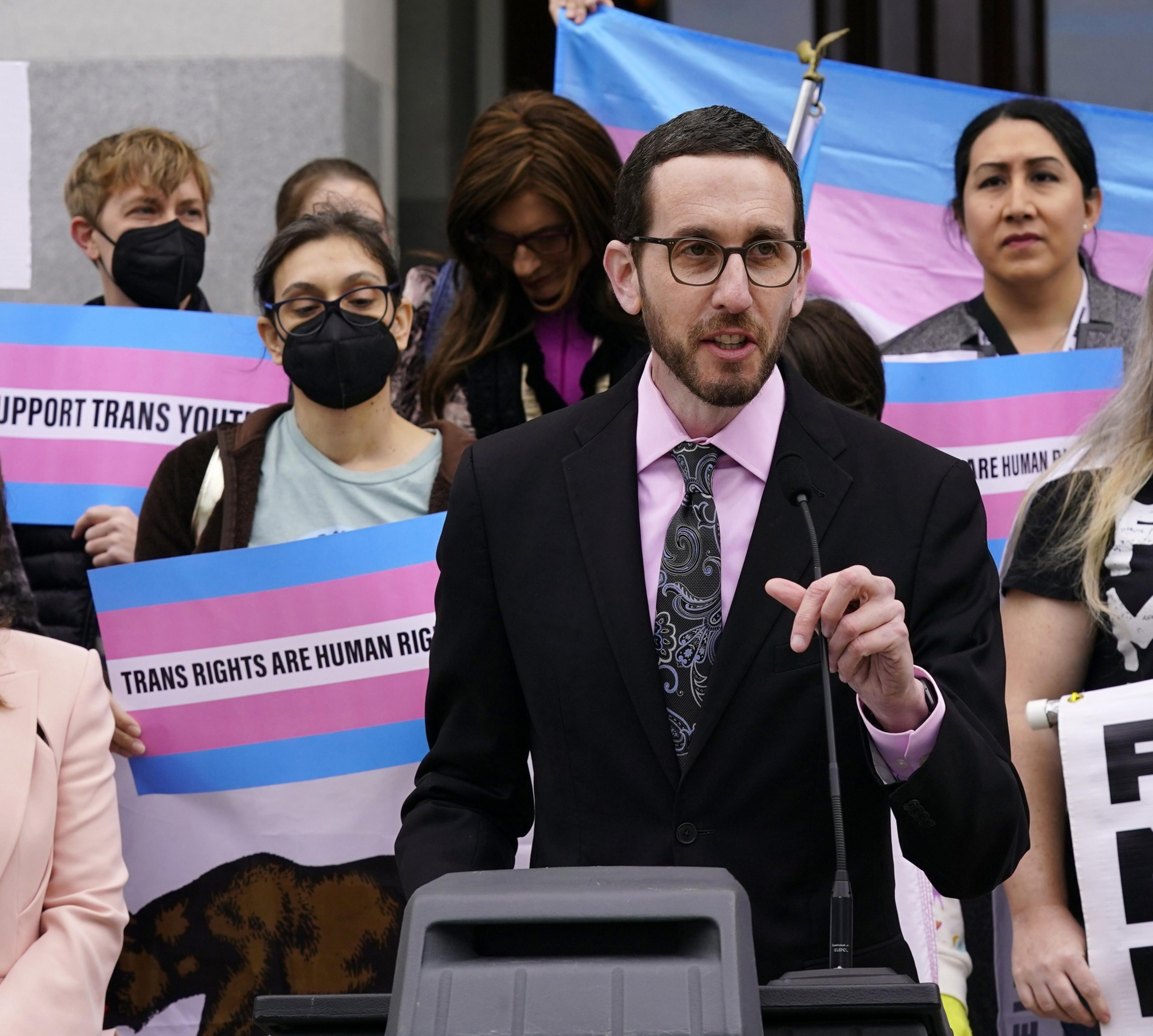 A person in a suit speaks at a podium with a crowd behind them holding pink and blue signs reading &quot;Trans Rights Are Human Rights.&quot;