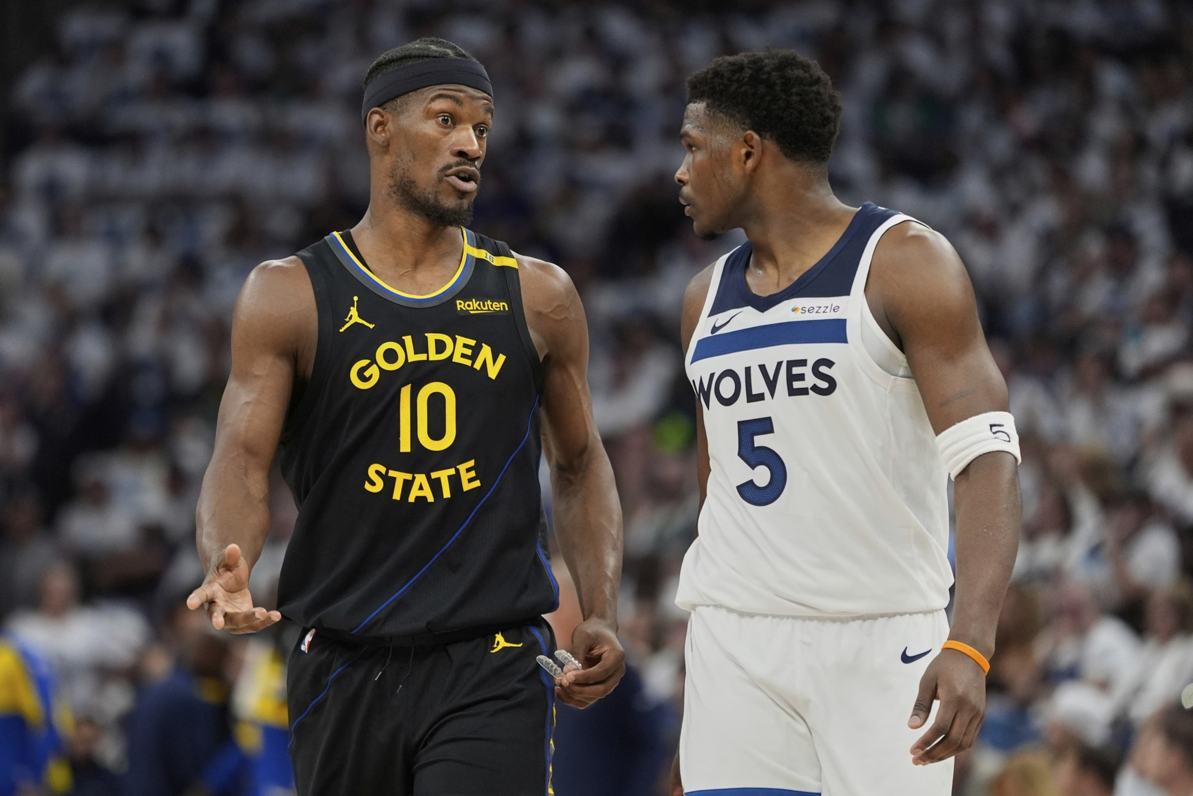 Two basketball players are on the court. One wears a black Golden State jersey, the other a white Wolves jersey. They're talking during a game.