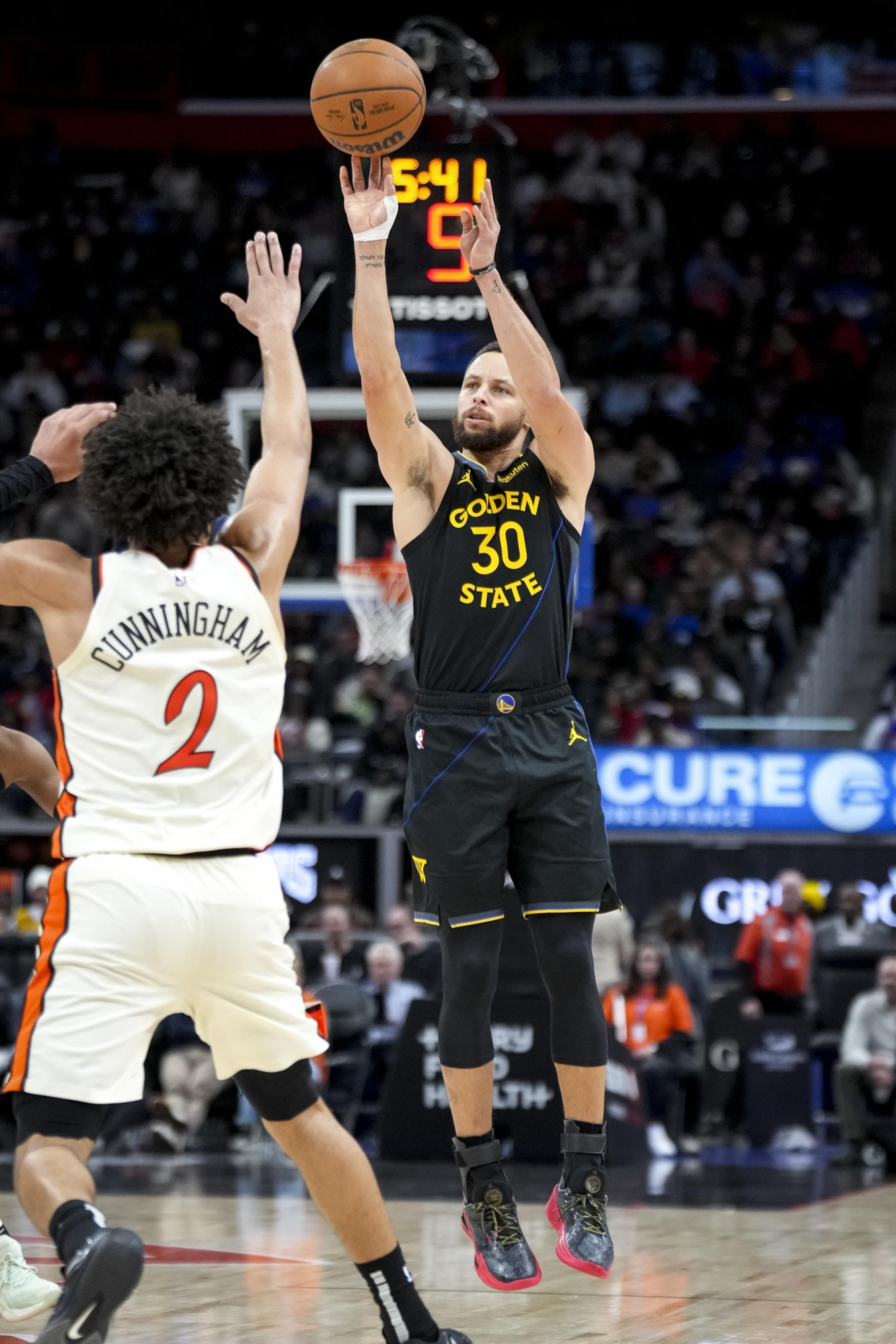 A basketball player in a black Golden State jersey shoots a ball mid-air while another player in a white jersey defends him. The crowd is visible in the background.