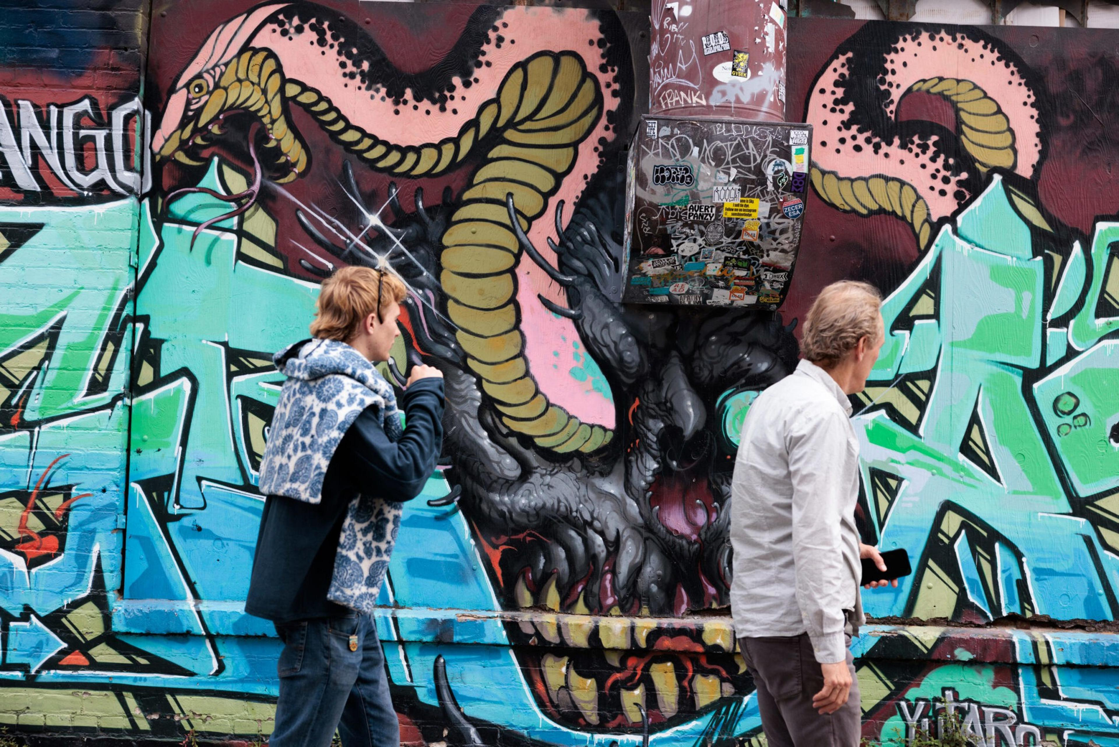 A large mural of a dark skull with yellow snake-like horns covers a wall, while two men walk past, one drinking from a cup.