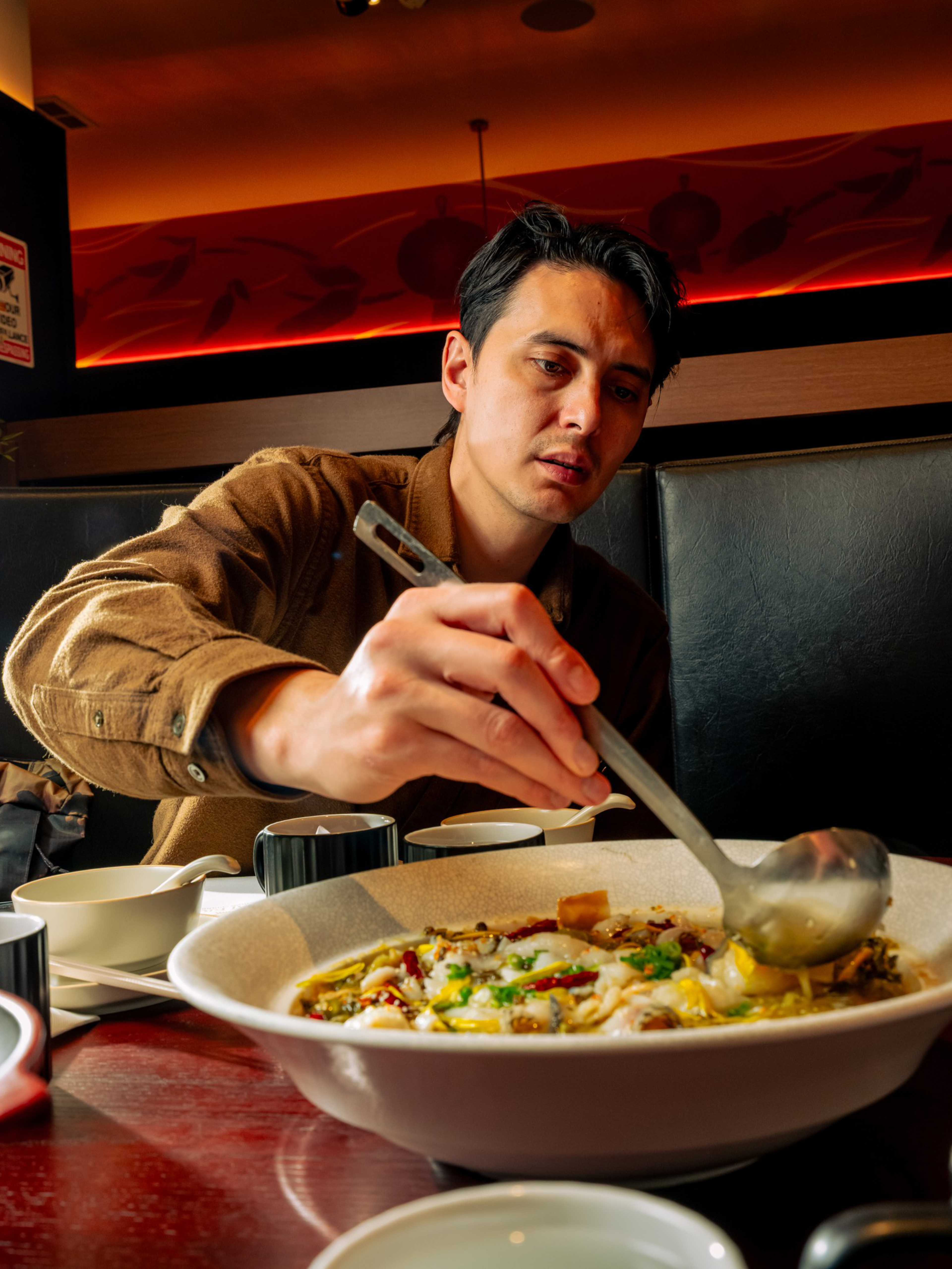 A man wearing a brown shirt is ladling soup from a large bowl at a dimly lit restaurant table with cups and spoons.