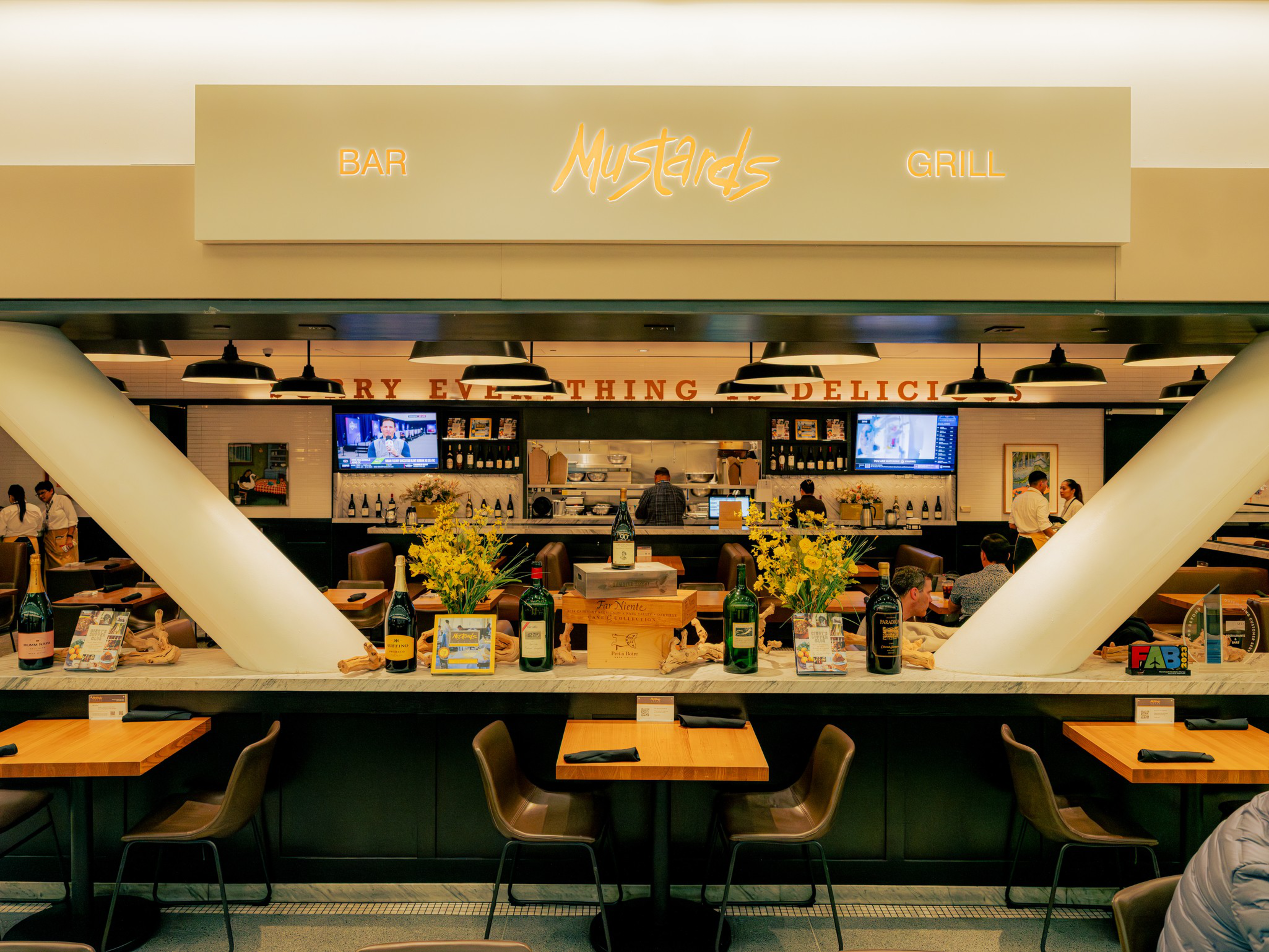 A bar and grill named Mustards features wooden tables with chairs, large white columns, bottles and flowers on the counter, and staff behind the bar.