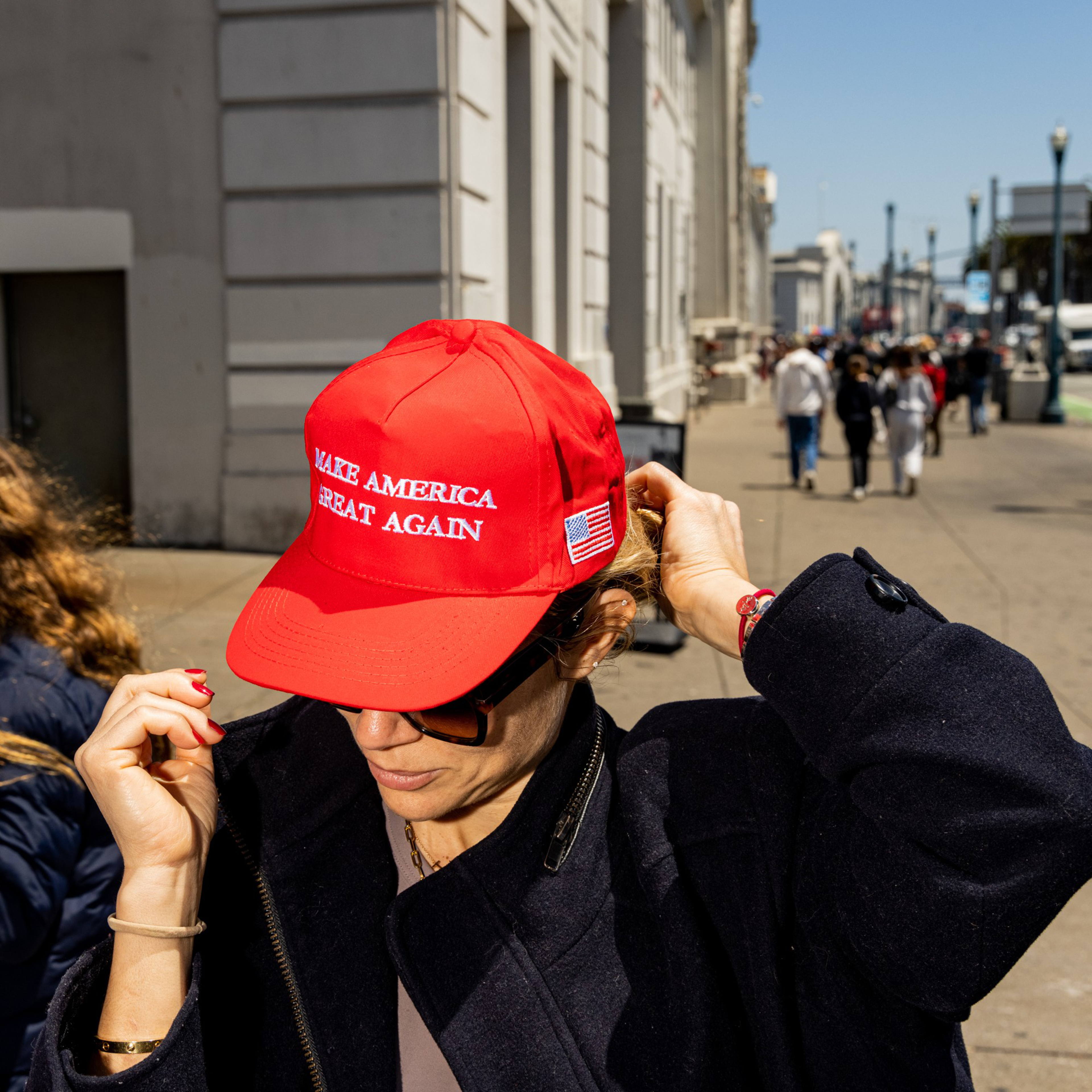 A person is adjusting their red &quot;Make America Great Again&quot; cap while wearing dark sunglasses and a navy coat, standing on a busy sidewalk with buildings in the background.
