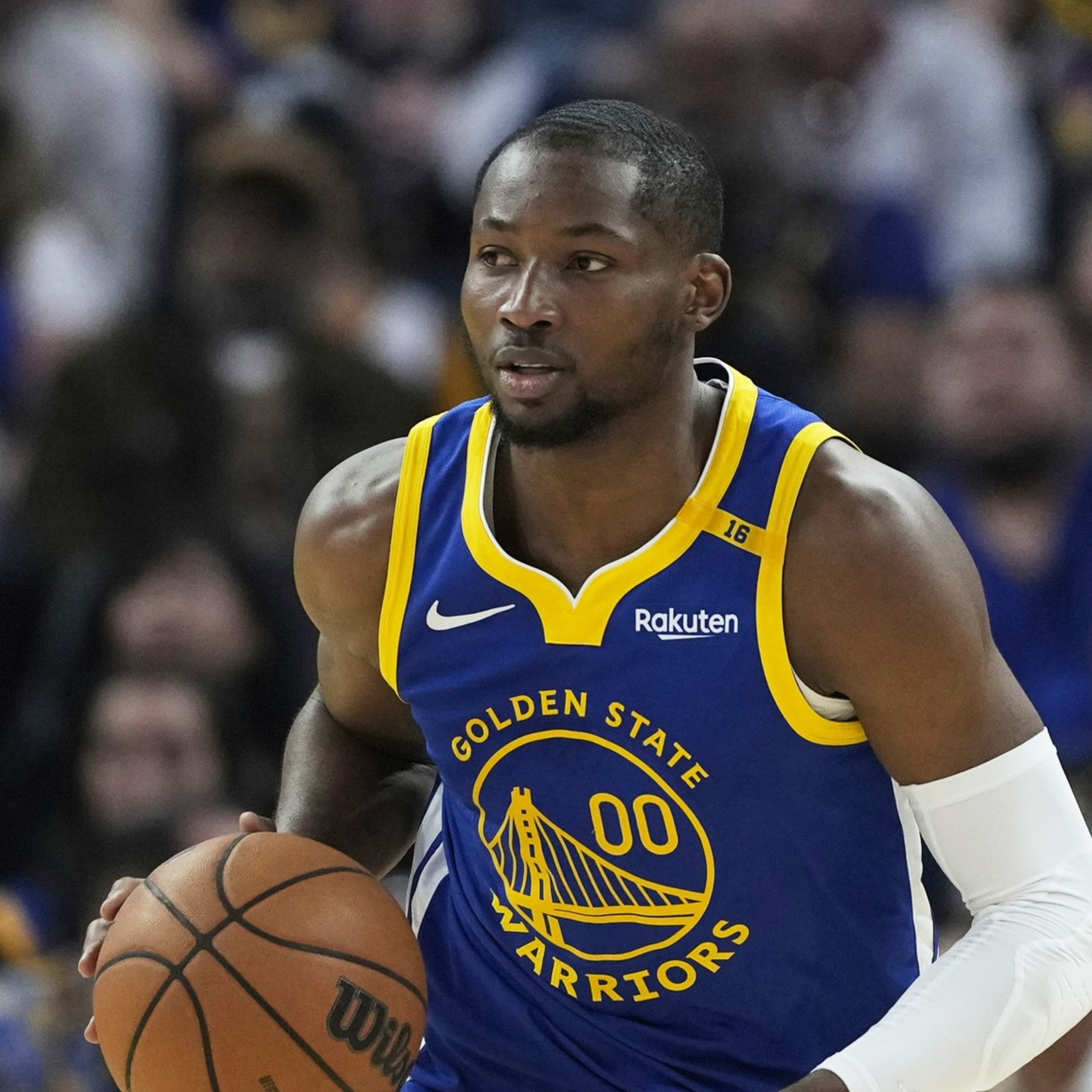 A basketball player in a Golden State Warriors jersey is dribbling the ball on the court. Spectators and an official are blurred in the background.