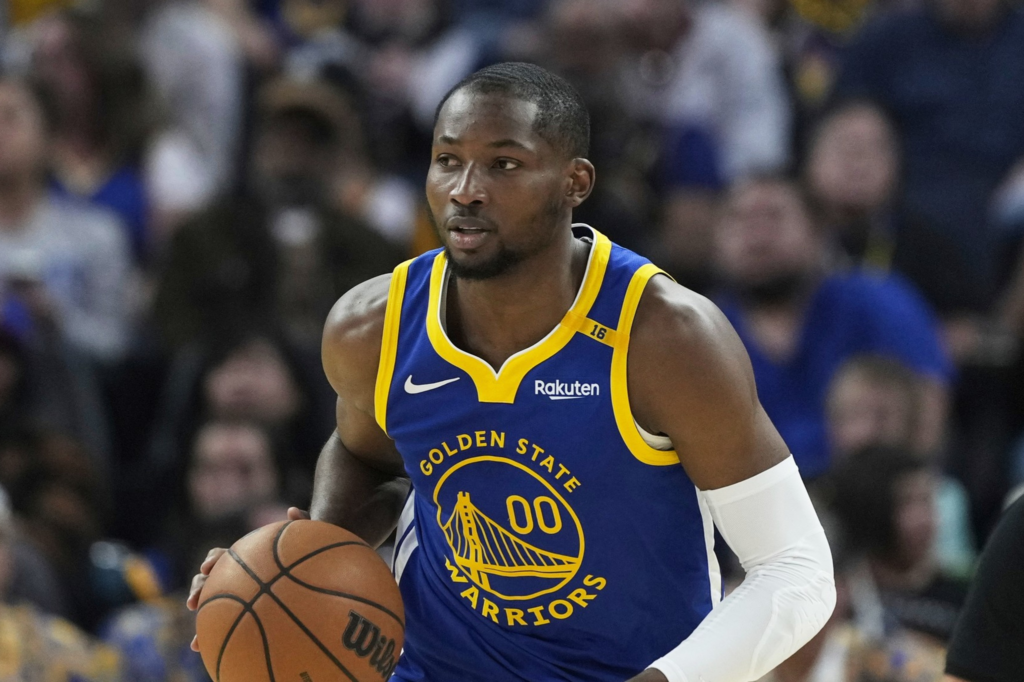 A basketball player in a Golden State Warriors jersey is dribbling the ball on the court. Spectators and an official are blurred in the background.