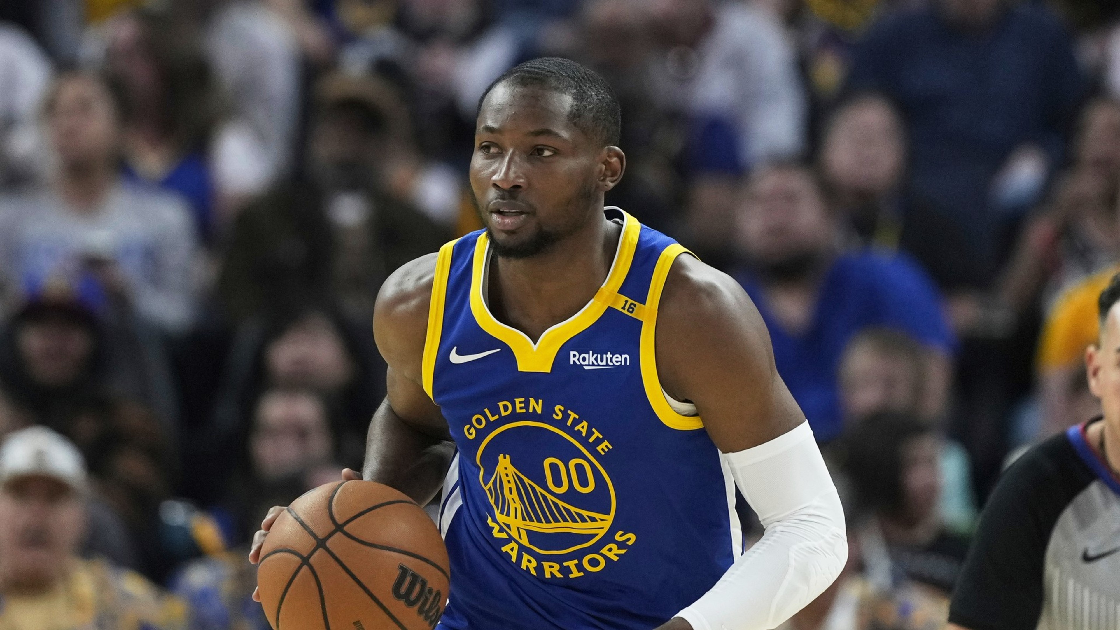 A basketball player in a Golden State Warriors jersey is dribbling the ball on the court. Spectators and an official are blurred in the background.
