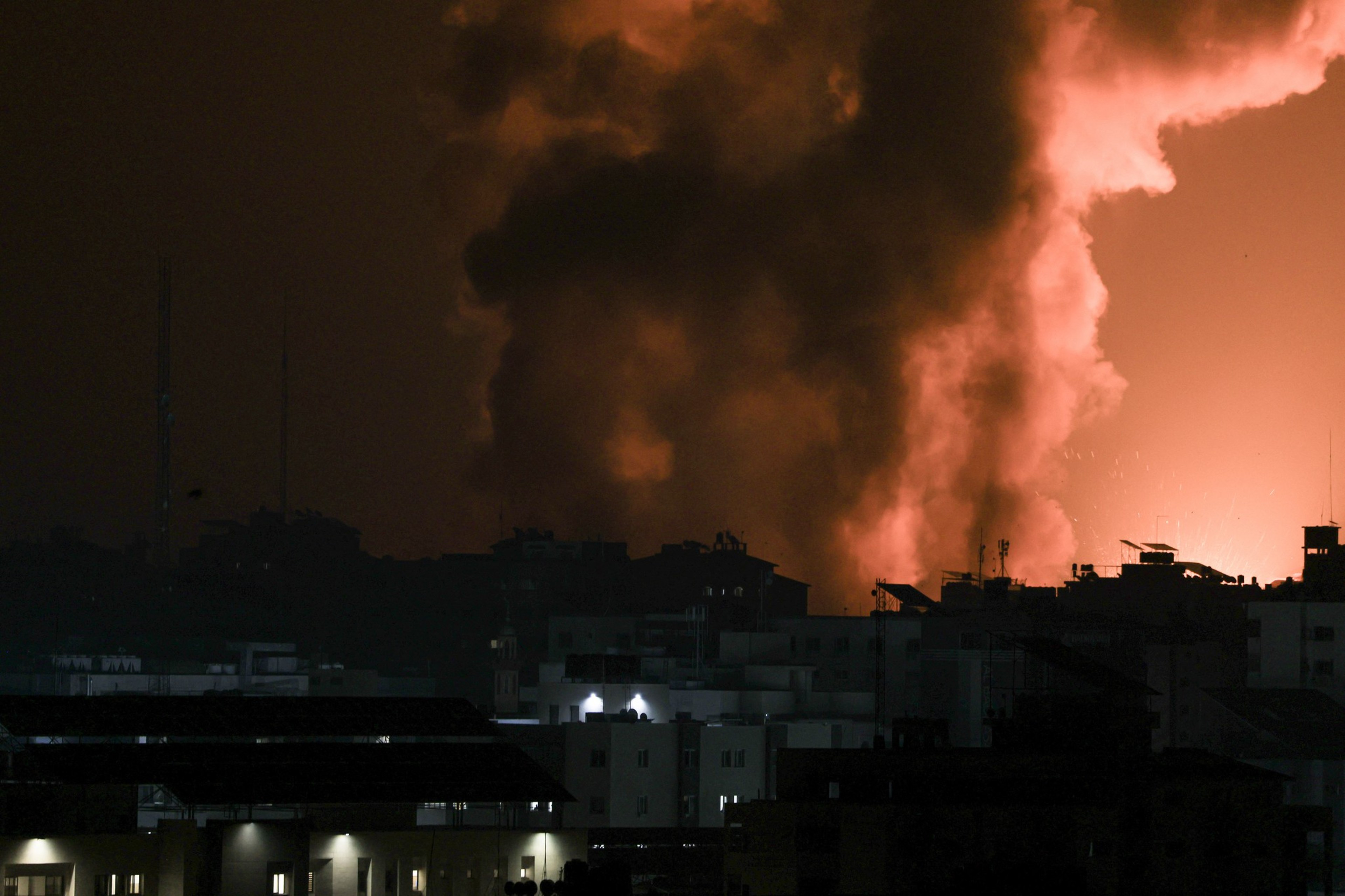 A city skyline at night is lit by a fiery glow and smoke in the background.