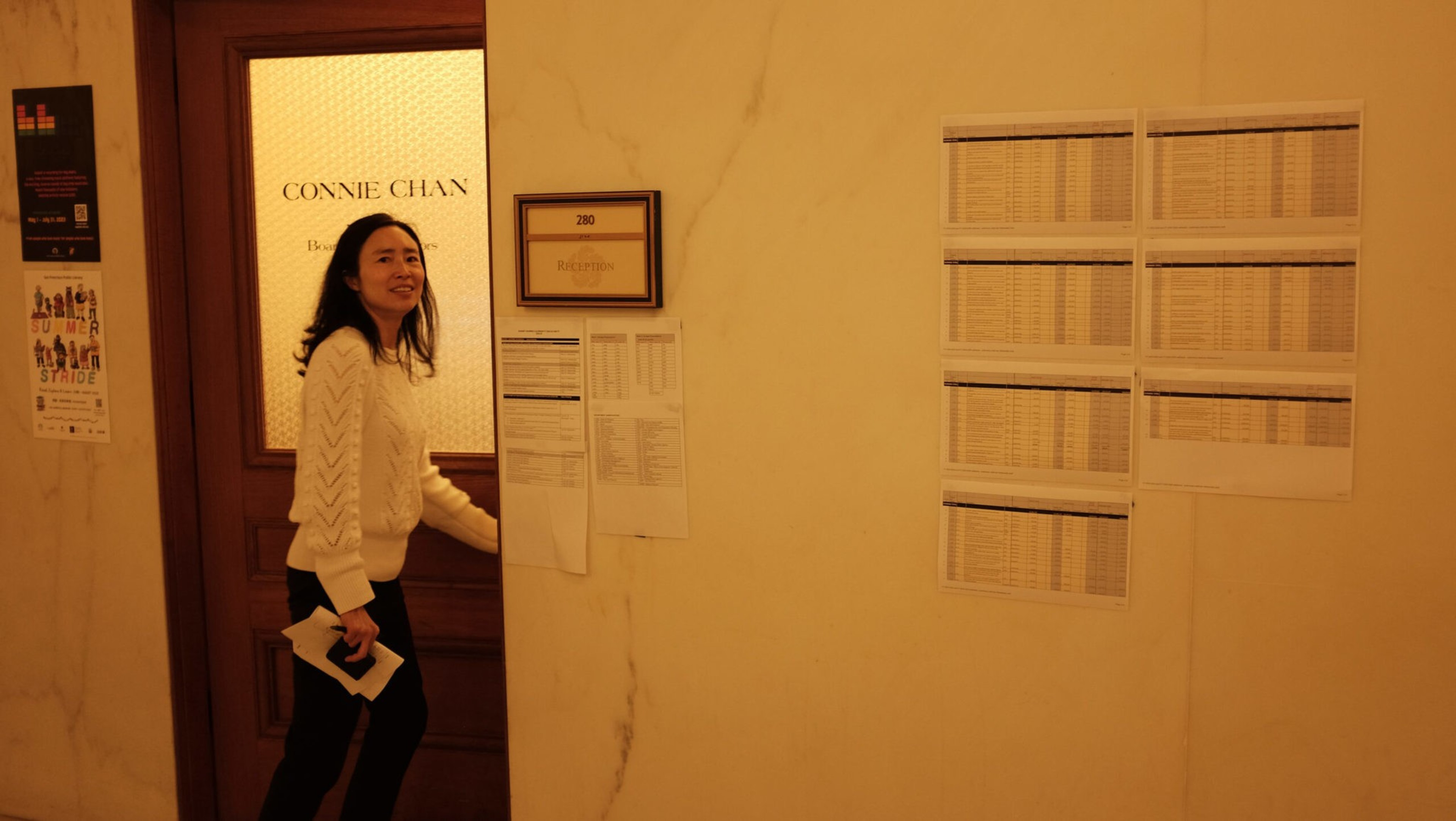 A woman in a white sweater holds a paper and opens a door labeled "CONNIE CHAN" with various schedules posted on the wall beside it.