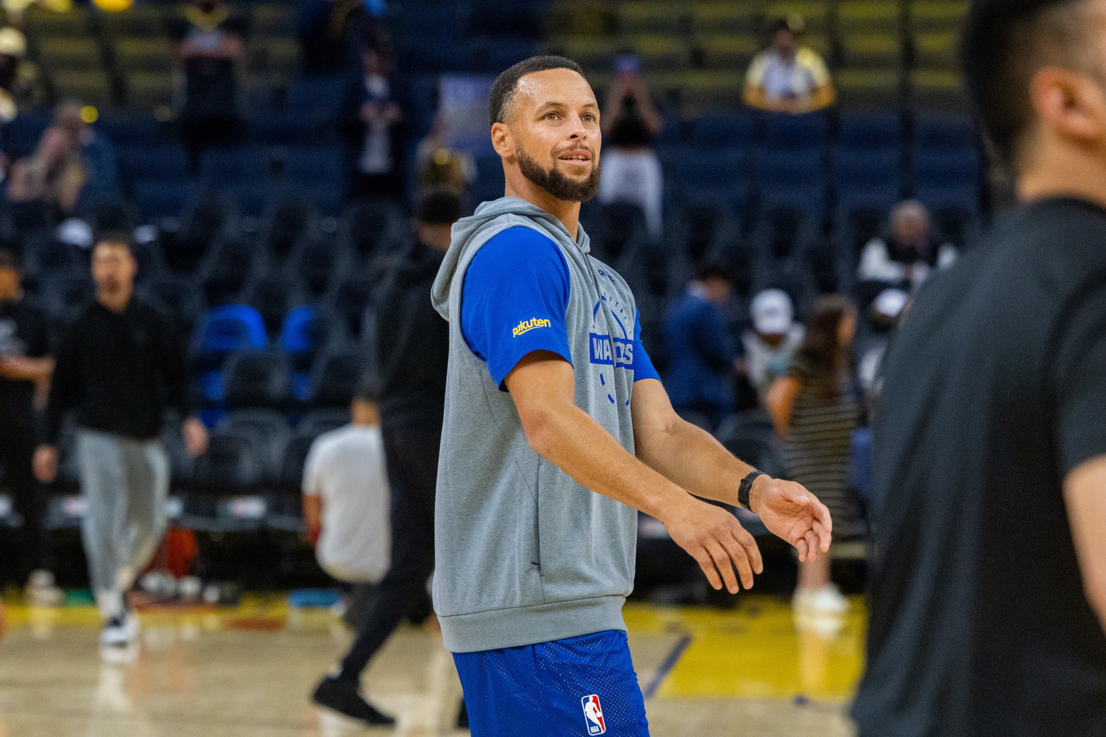 A basketball player in a blue and gray Golden State Warriors warm-up outfit is standing on the court, preparing to shoot or pass the ball.