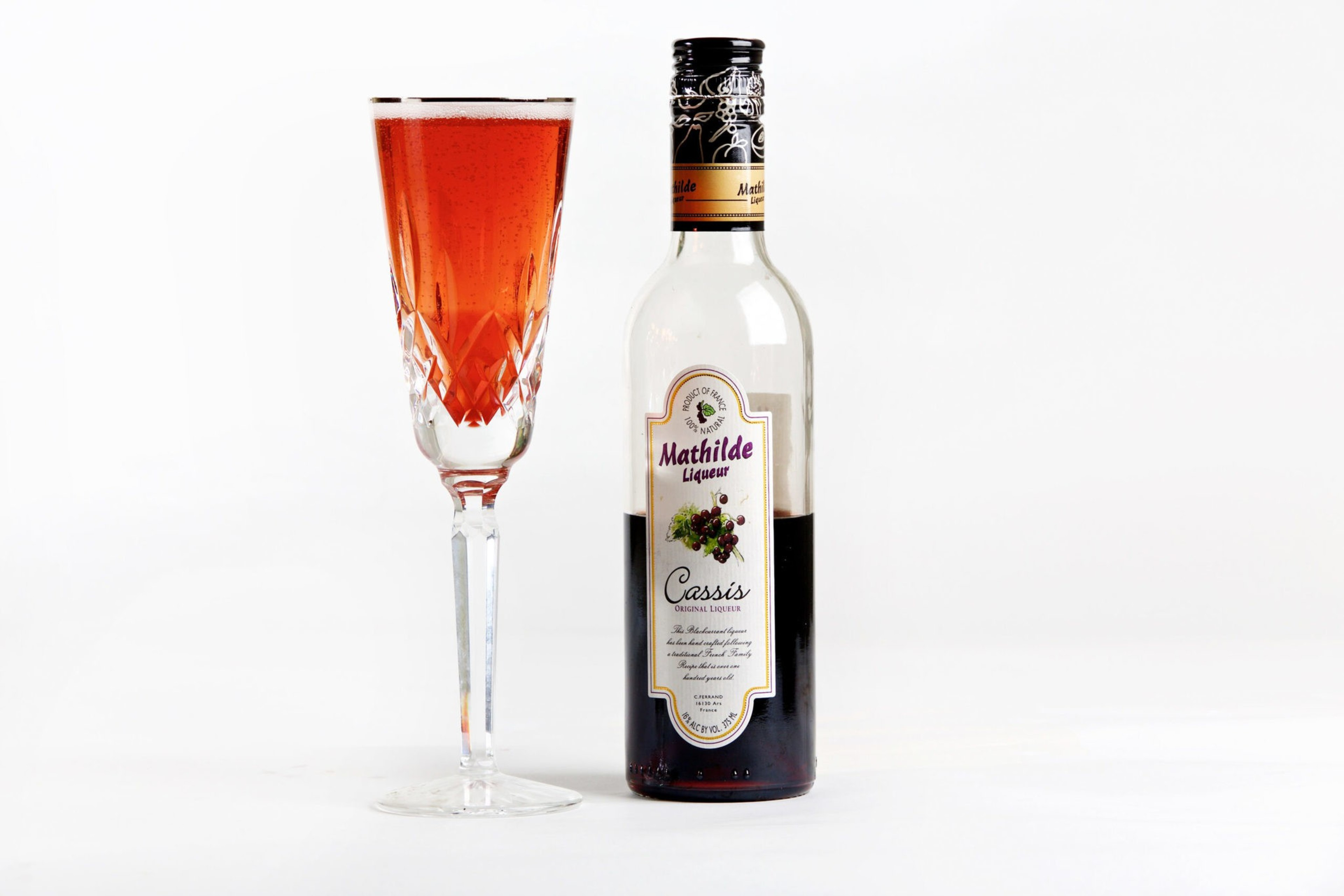 A tall, clear glass holds a red liquid next to a partially full bottle ...