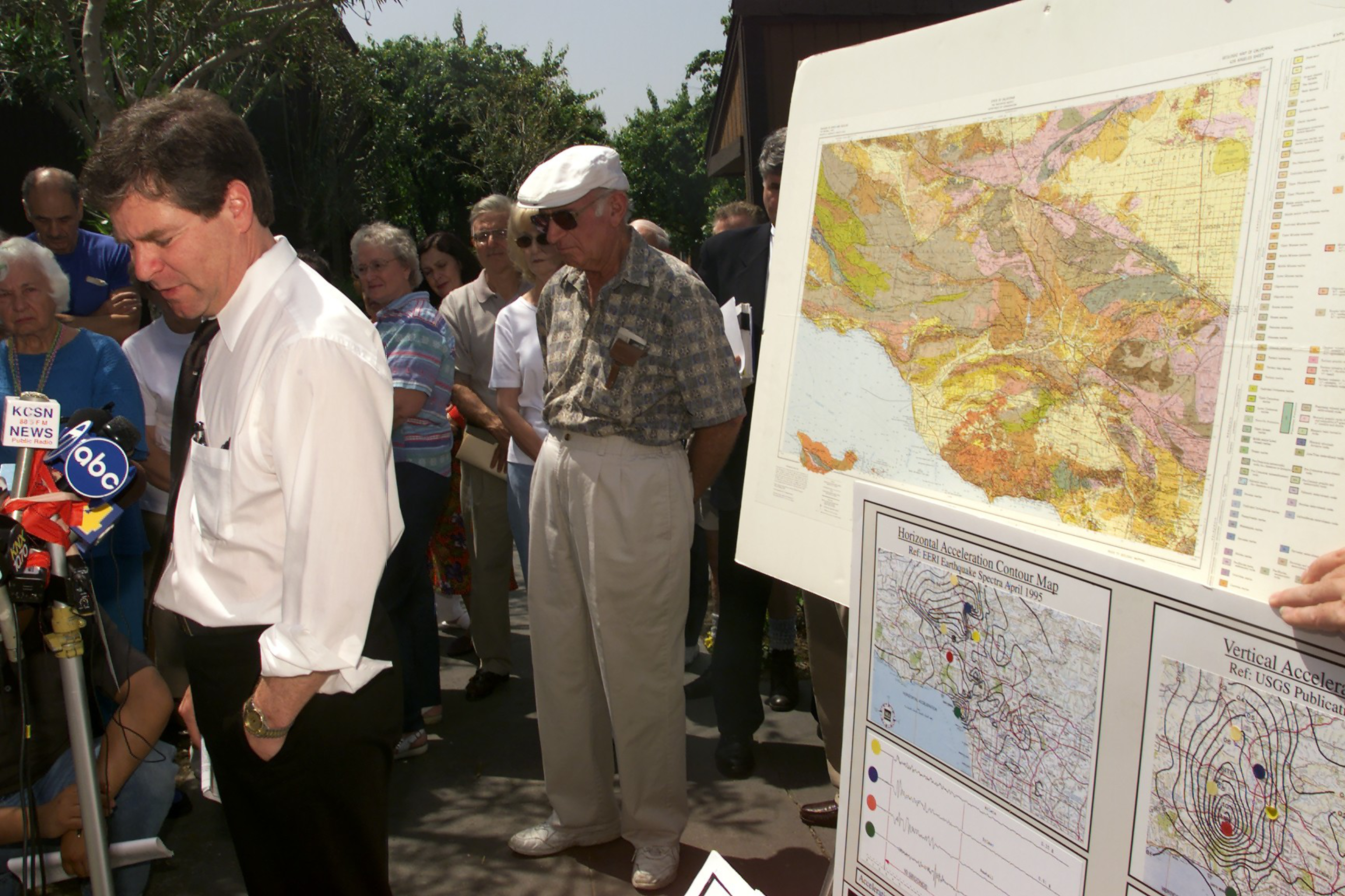 A man in a white shirt speaks near microphones while a crowd listens and geological maps are displayed on a board nearby.