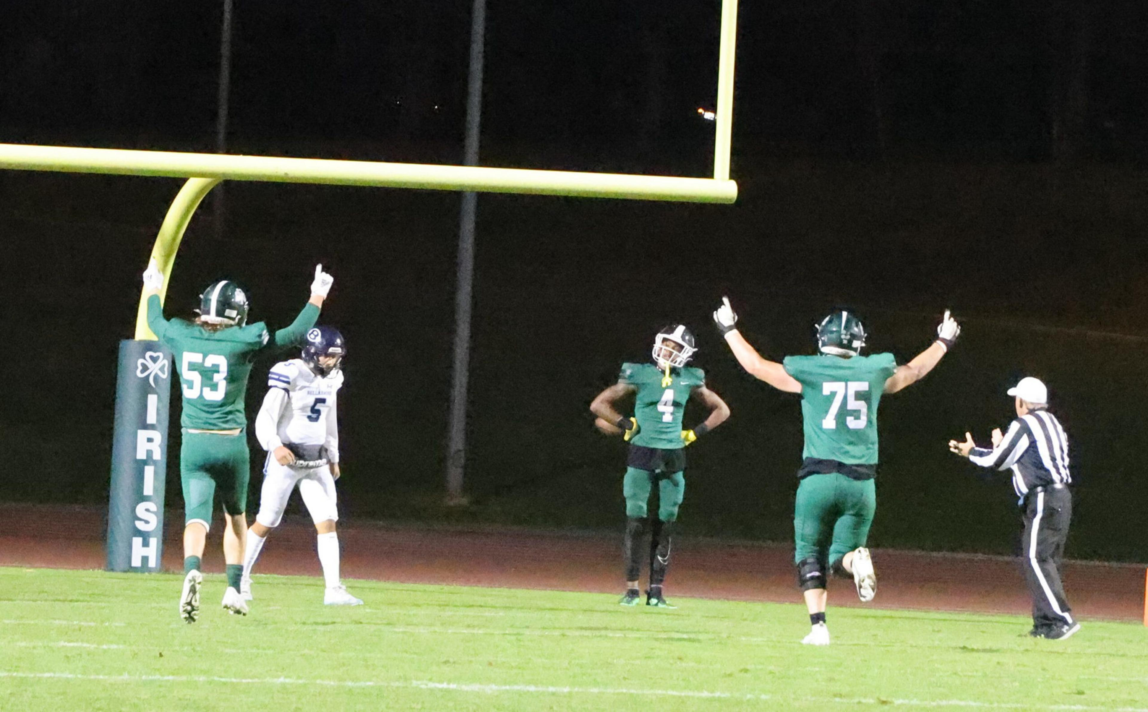 Three football players in green uniforms celebrate a touchdown near the goalpost while a referee signals and a player in white looks on.