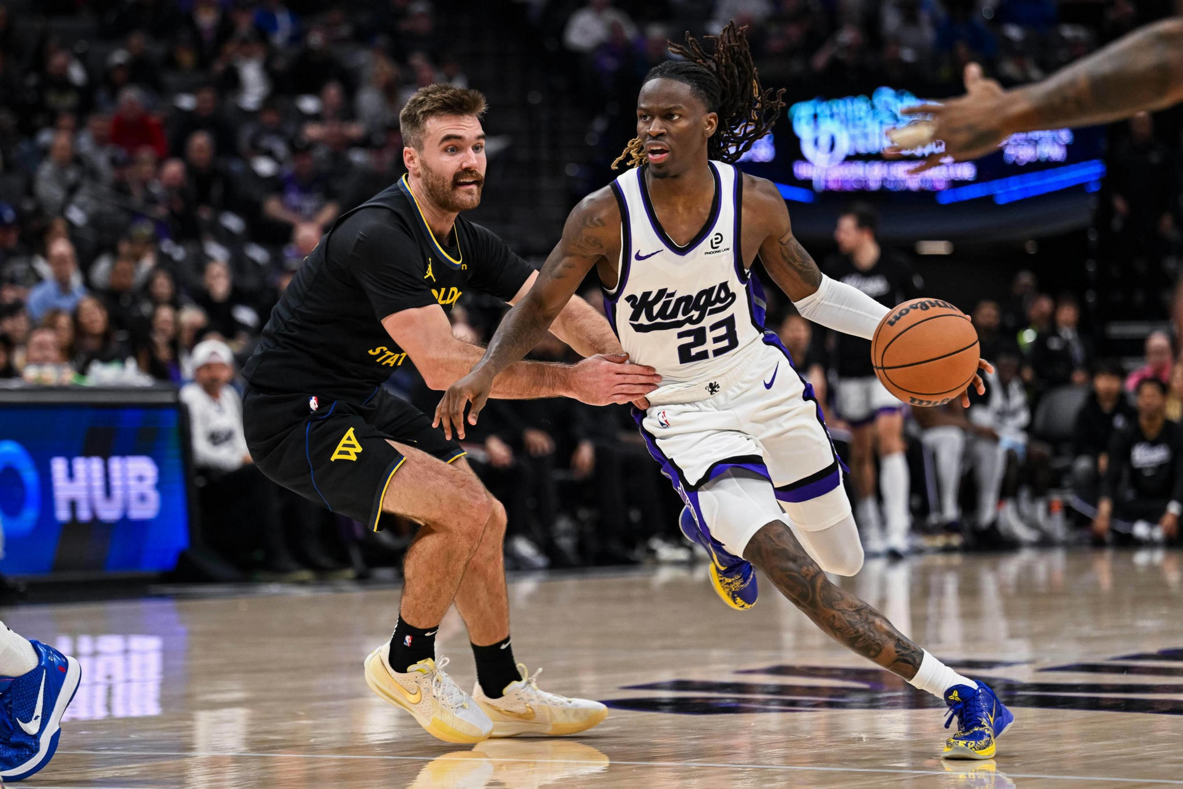 A basketball player in a white Kings uniform dribbles past a defender in a black Golden State uniform during a game.