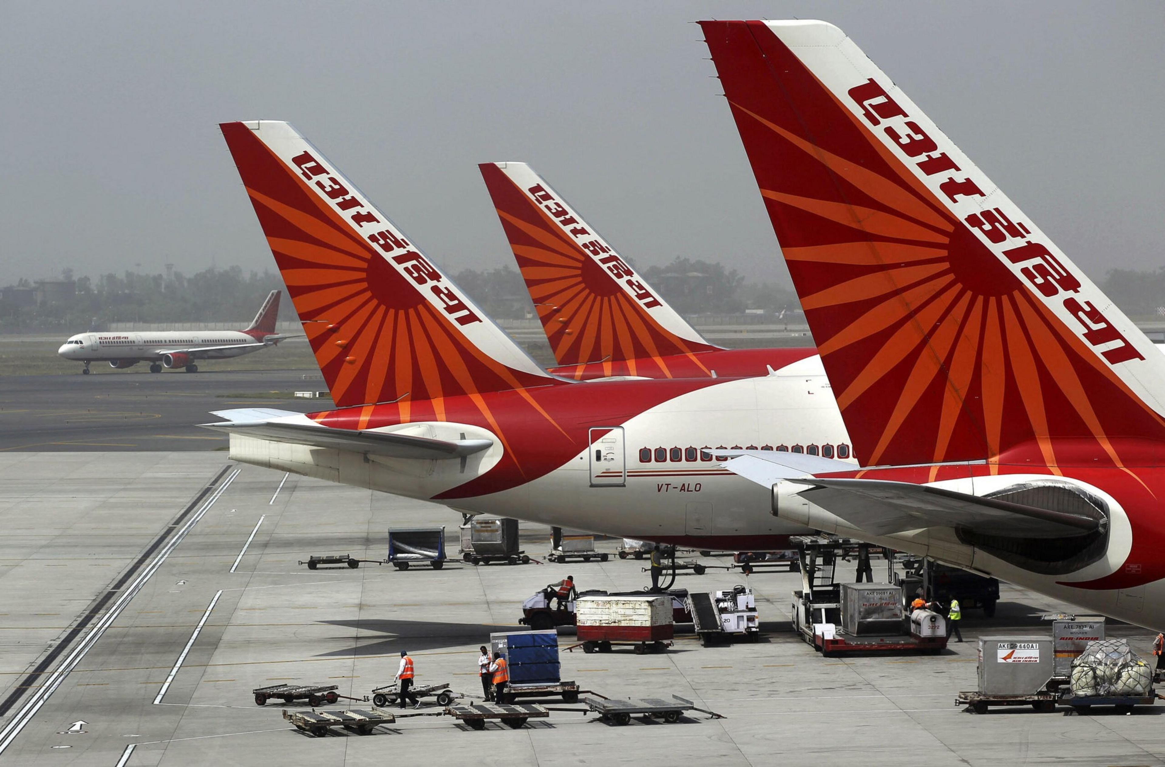 Three Air India planes with red and orange sunburst tails are parked on the tarmac while ground crew manage baggage and cargo near them.
