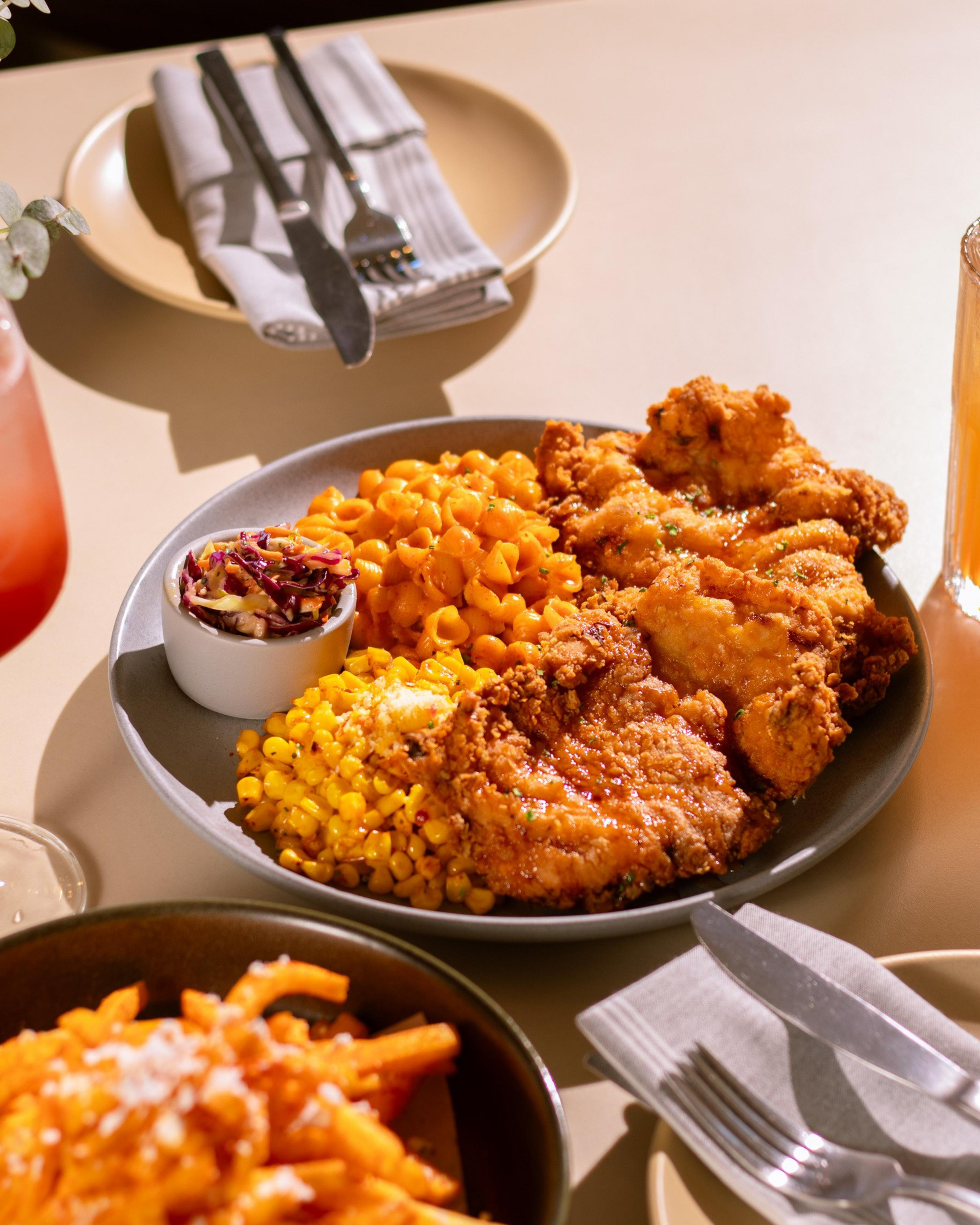 Crispy fried chicken served with macaroni and cheese, buttered corn, and a small cup of coleslaw on a gray plate. Silverware and drinks nearby.