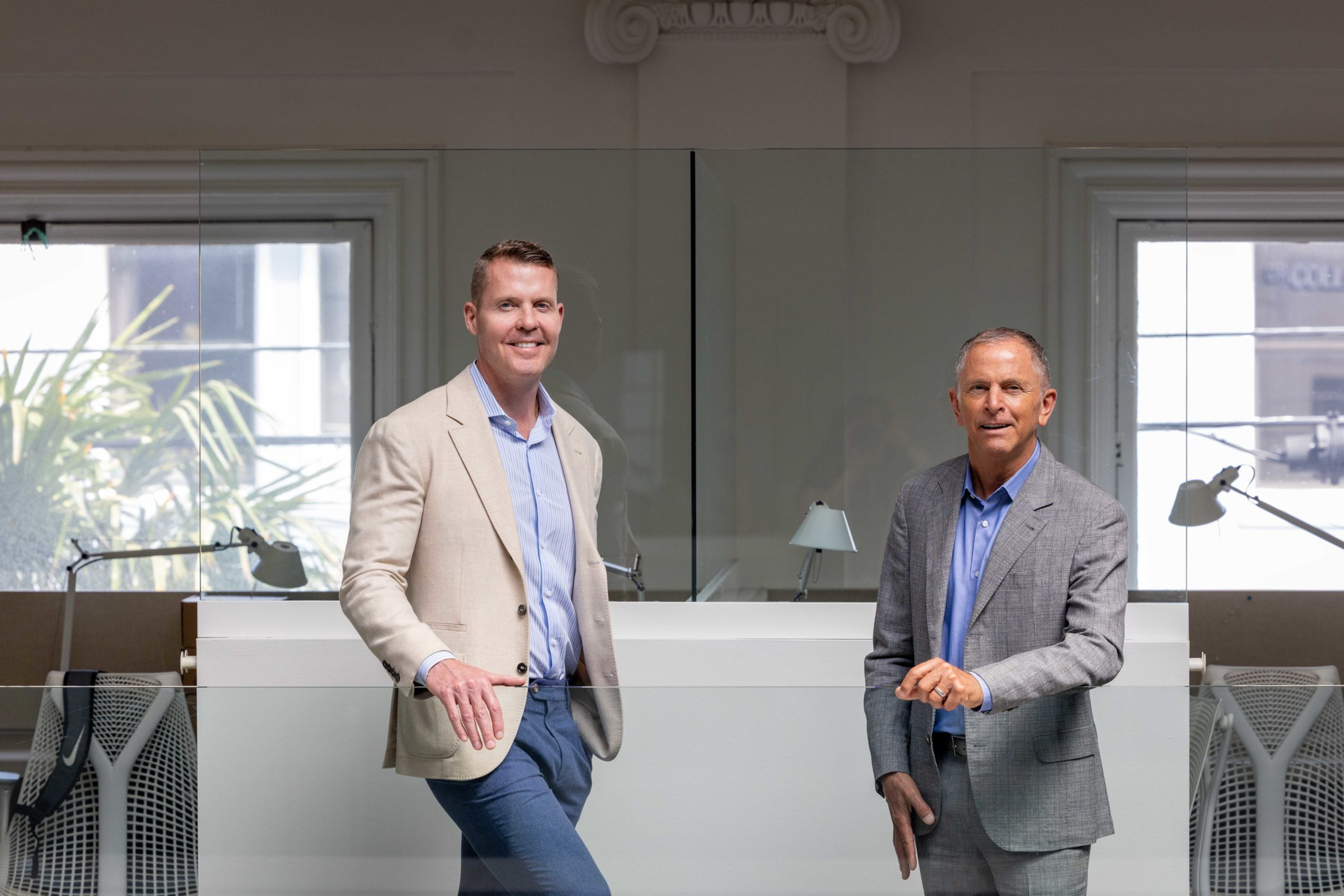 Two men in business attire stand in a modern office with large windows, glass partitions, and desk lamps behind them.