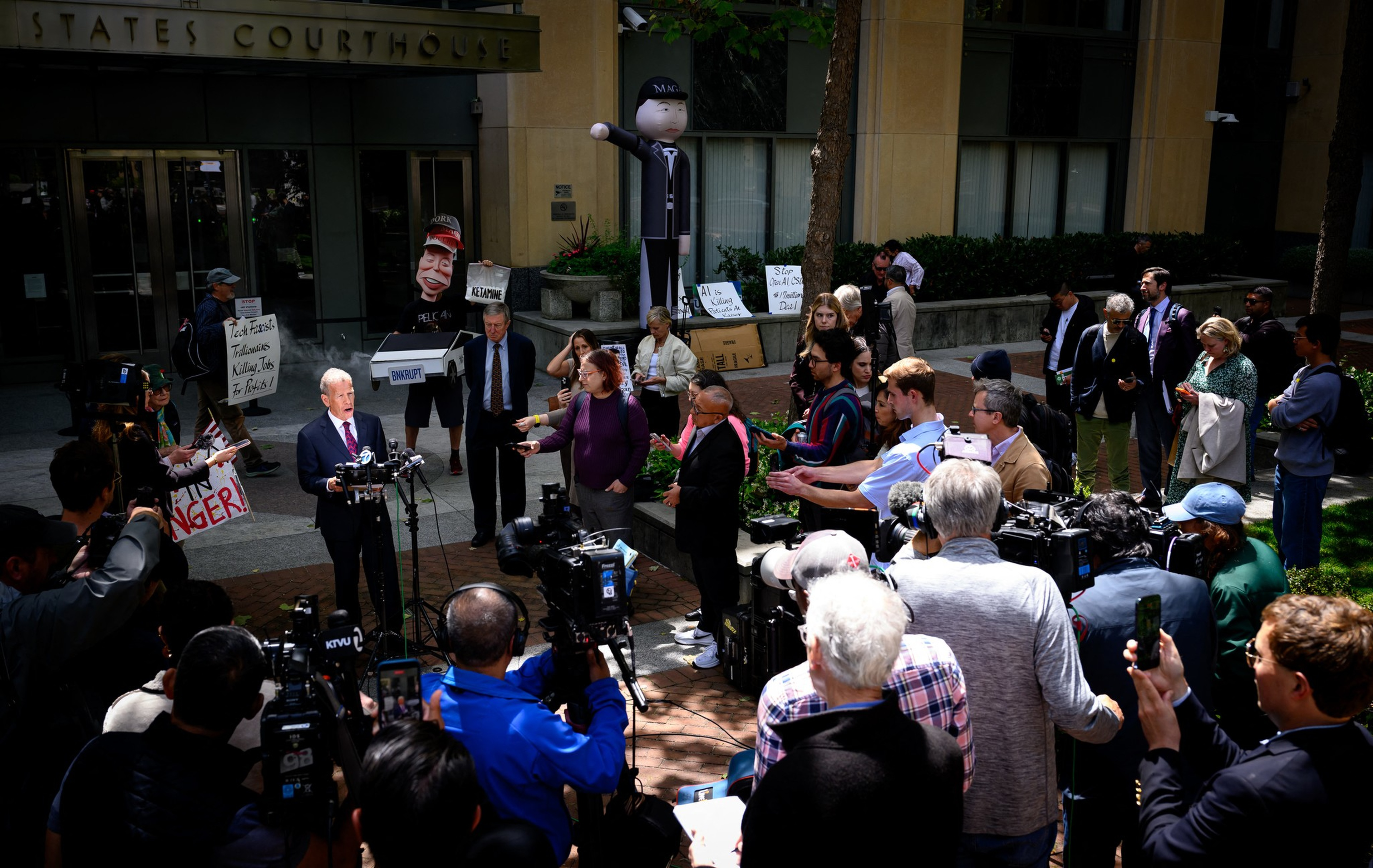 A man in a suit speaks at a press conference outside a courthouse, surrounded by reporters, cameras, and protest signs with caricatures and slogans.
